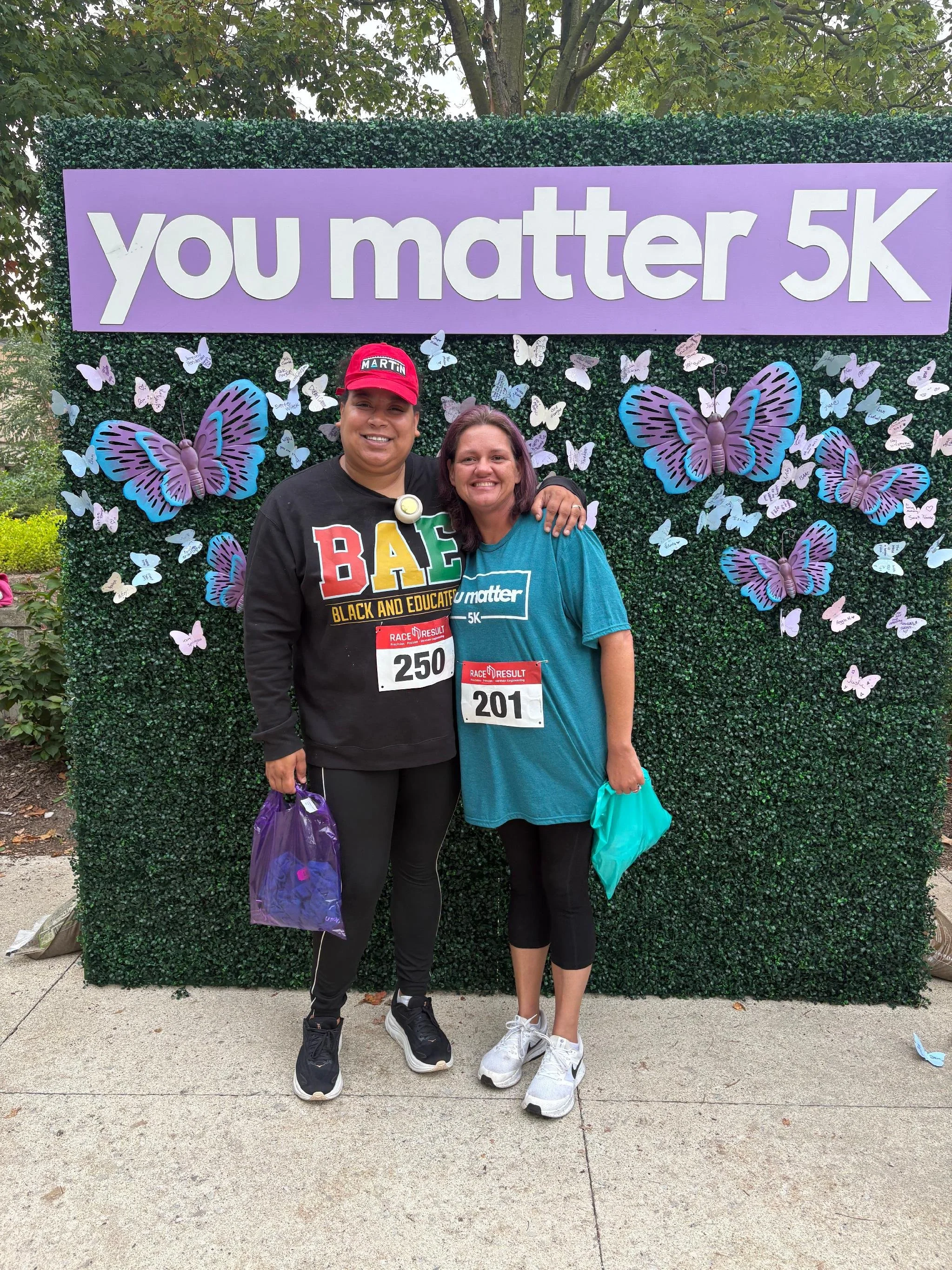 Two women standing together smiling in front of a purple sign that says "you matter 5K" with butterfly decorations on a green hedge background. One woman is wearing a black "B.A.E." sweatshirt and a red cap, the other is wearing a teal T-shirt with t