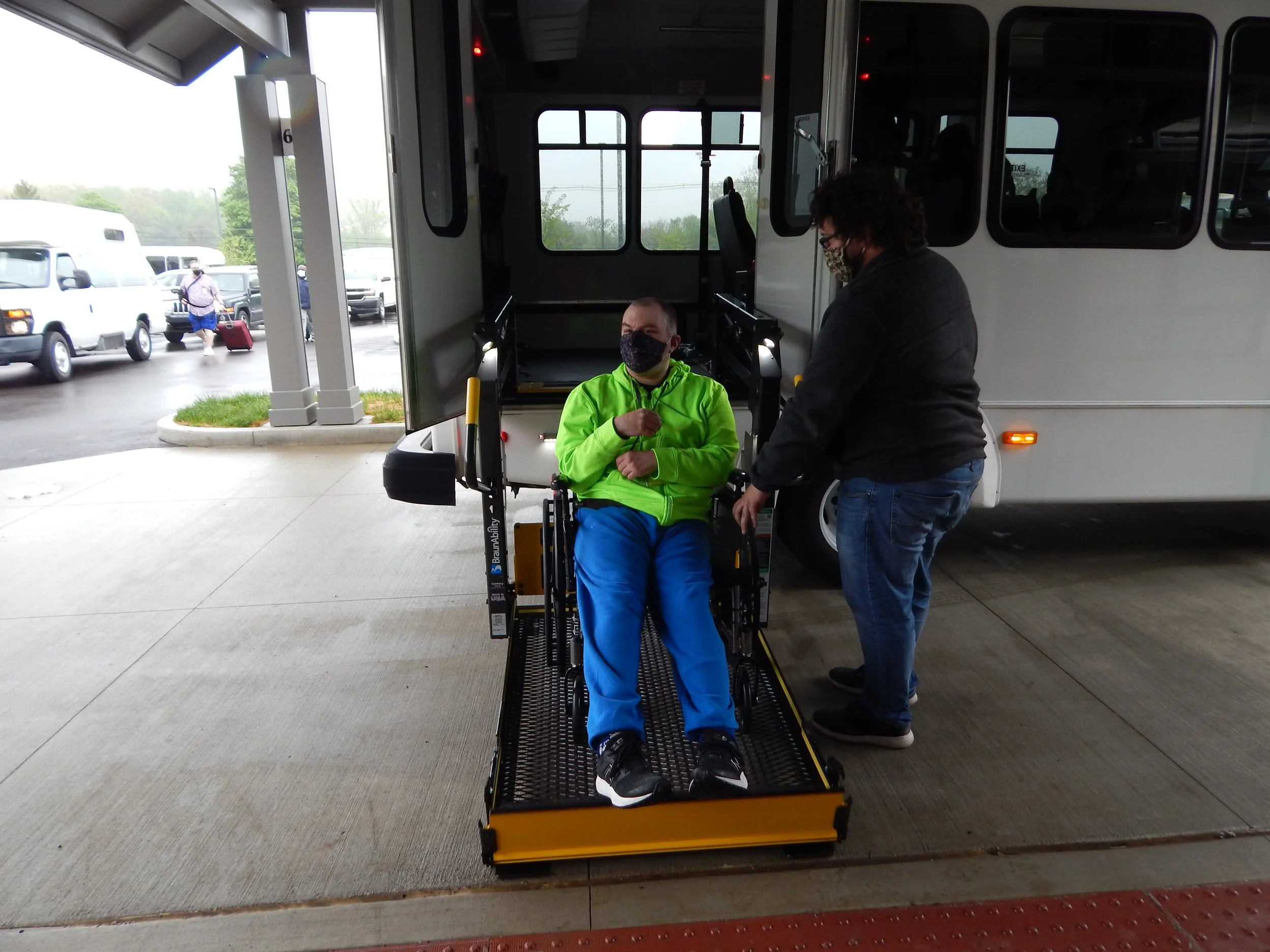 A person in a bright green jacket and blue pants is sitting in a wheelchair at a bus station, while another person standing nearby appears to be assisting. Both individuals are wearing face masks and are positioned on a wheelchair ramp outside a bus.