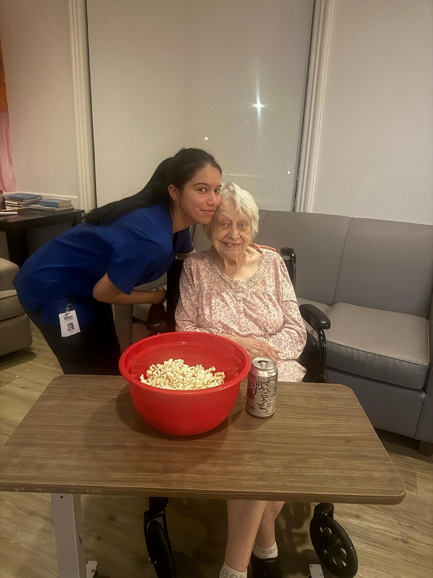 A young woman in a blue nurse uniform is leaning over an elderly woman sitting in a wheelchair. They are in a room with beige walls and a gray sofa. On the table in front of them, there is a large red bowl filled with popcorn, a soda can, and a soda 