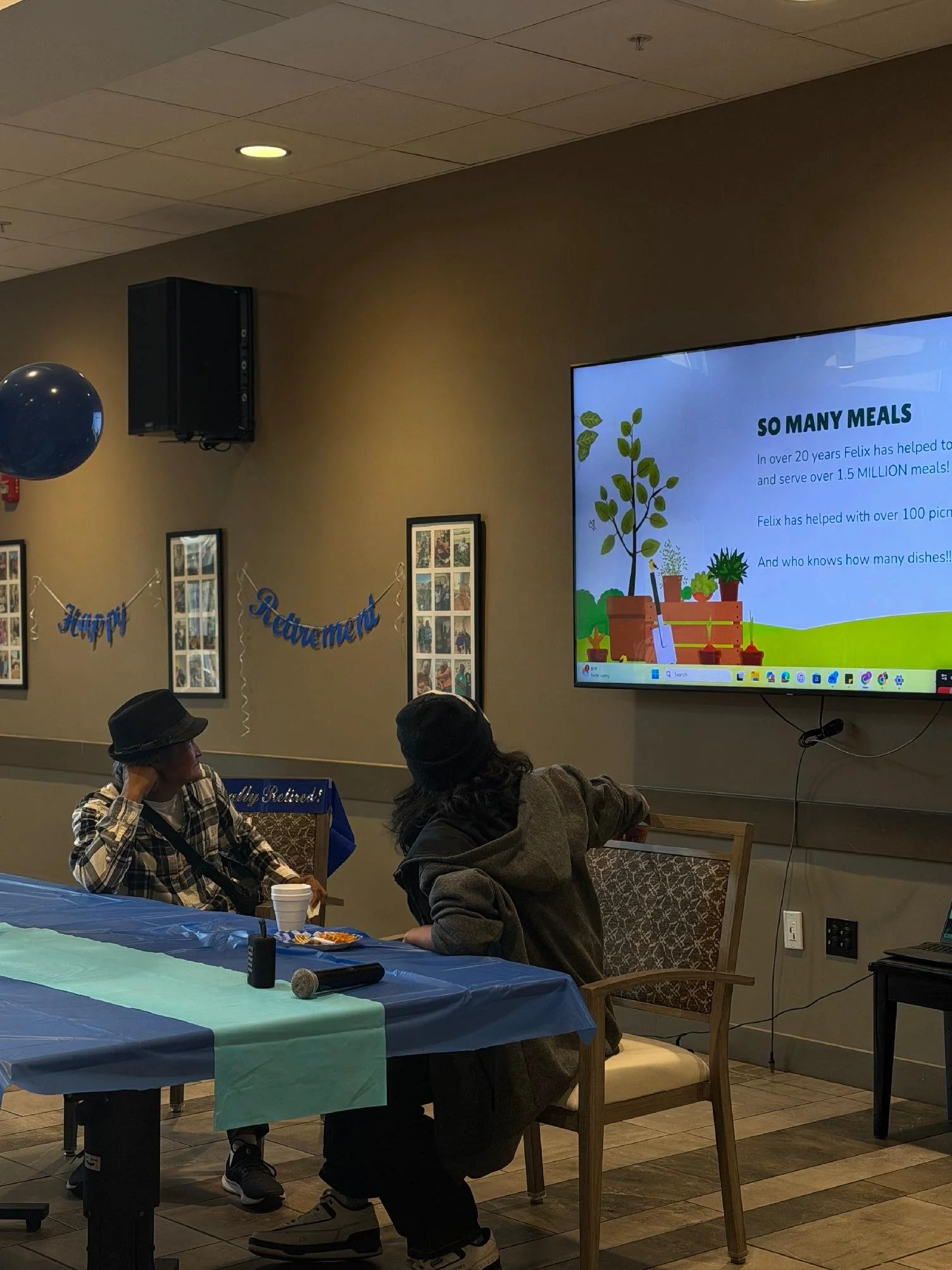 Indoor scene at a celebration with a large screen displaying a presentation, a table with teal tablecloth, balloons, people sitting and standing, and decorations including a 'Happy Retirement' banner.