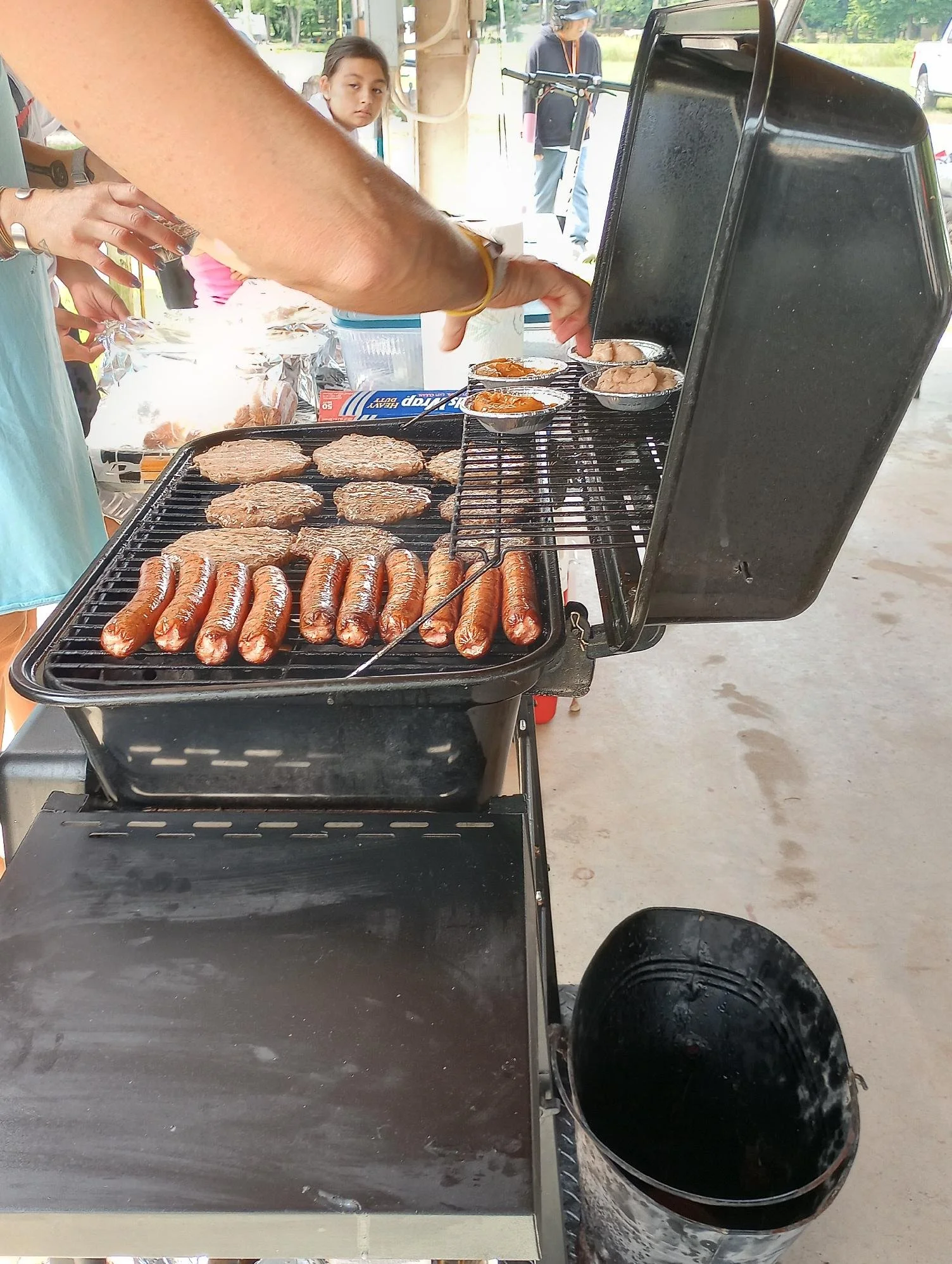A barbecue grill with hot dogs and hamburger patties cooking, with a person placing food into aluminum trays on the grill, and several people in the background at an outdoor gathering.