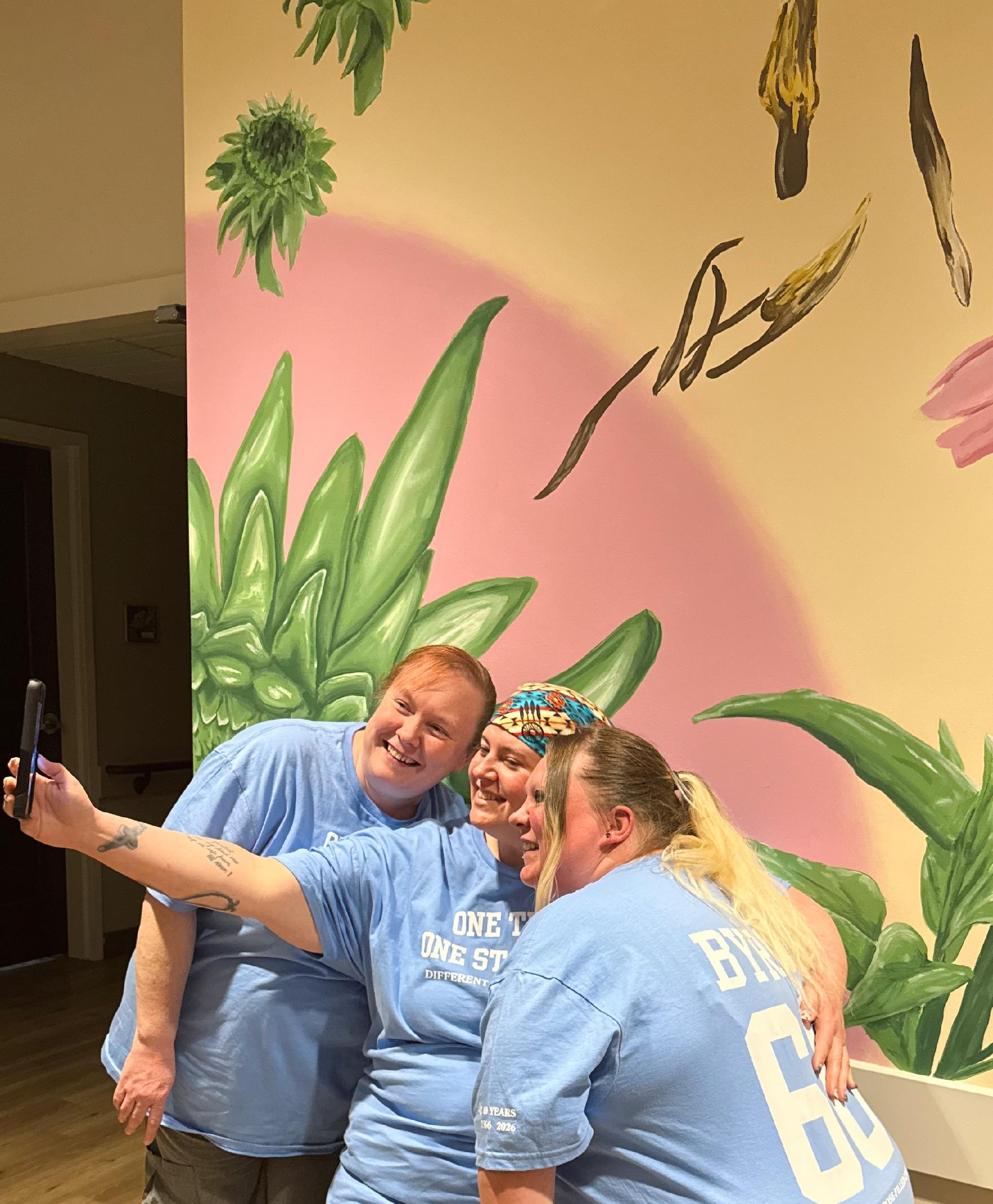 A group of people taking a selfie, smiling and hugging, in front of a large wall painting of green plants and pink flowers.