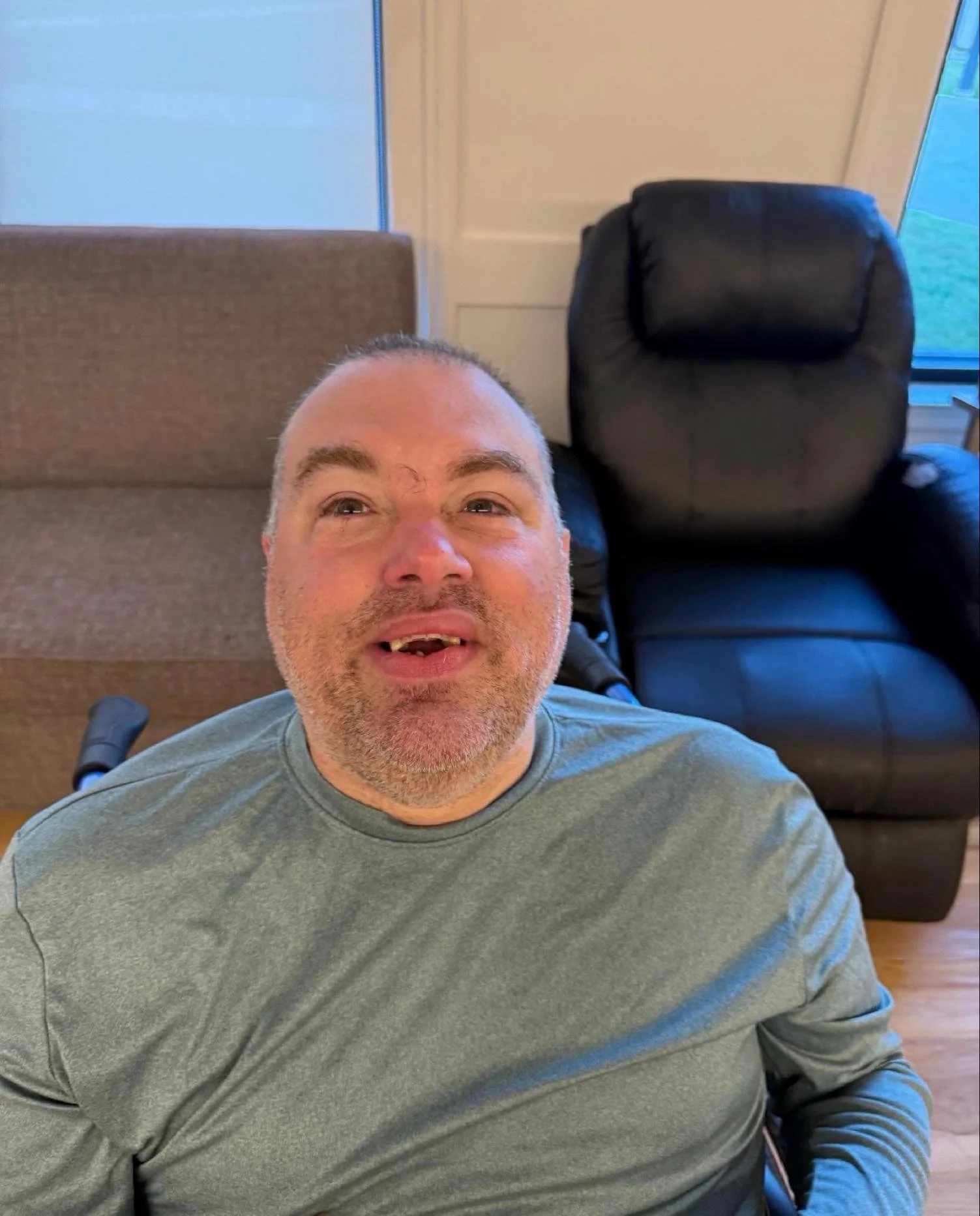 A man with short gray hair and a gray shirt, taking a selfie indoors, sitting on a black recliner chair near a brown sofa and a door with a window.