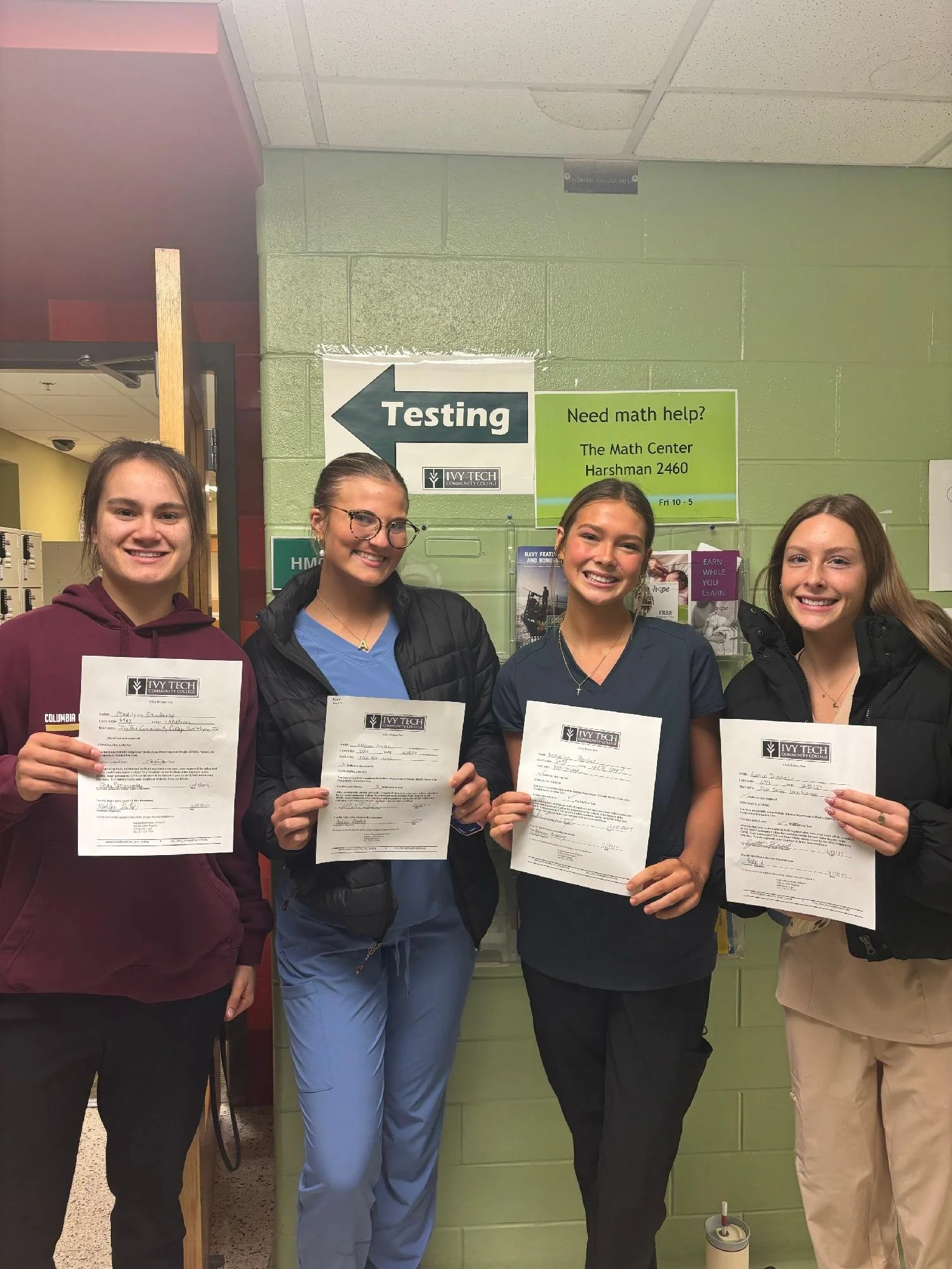 Four young women standing together indoors, holding papers, smiling for the camera. Behind them is a sign indicating testing direction and a green wall with additional informational signs.