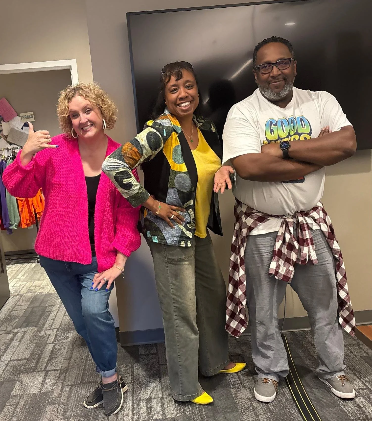 Four people standing in a room, smiling at the camera. The person on the left is a woman with curly blonde hair, wearing a bright pink sweater and blue jeans. Next to her is a woman with dark hair, wearing a colorful jacket, yellow shirt, and gray pa