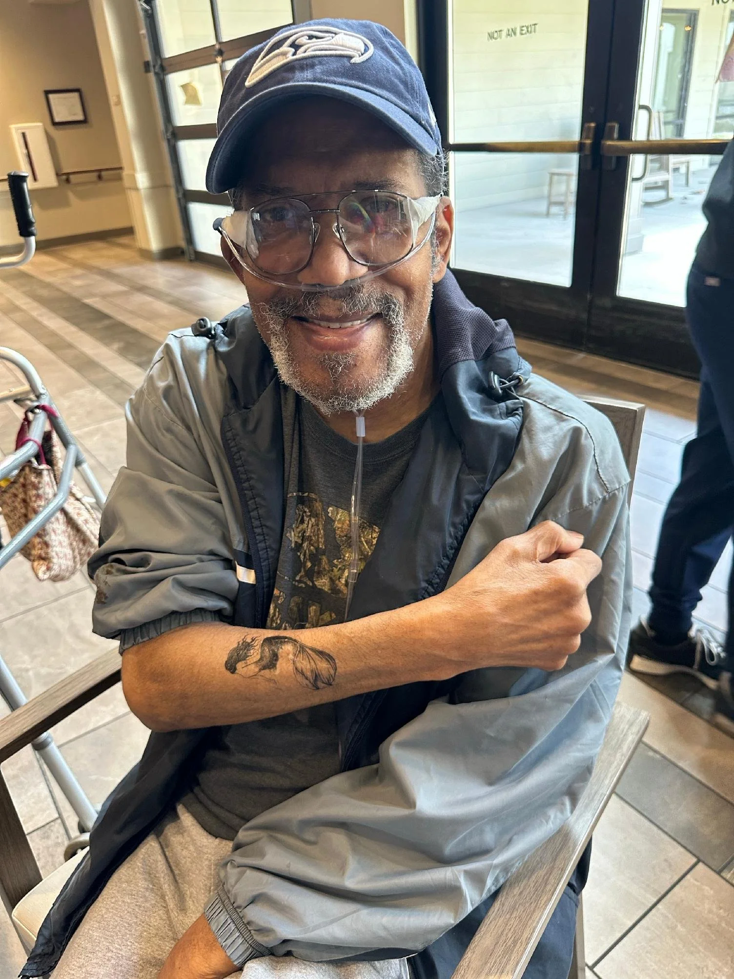 An elderly man with gray hair and glasses wearing a navy blue Seattle Seahawks baseball cap with a gray jacket, sitting at a table inside a building near glass doors, smiling, with a tattoo of a horse on his left forearm.