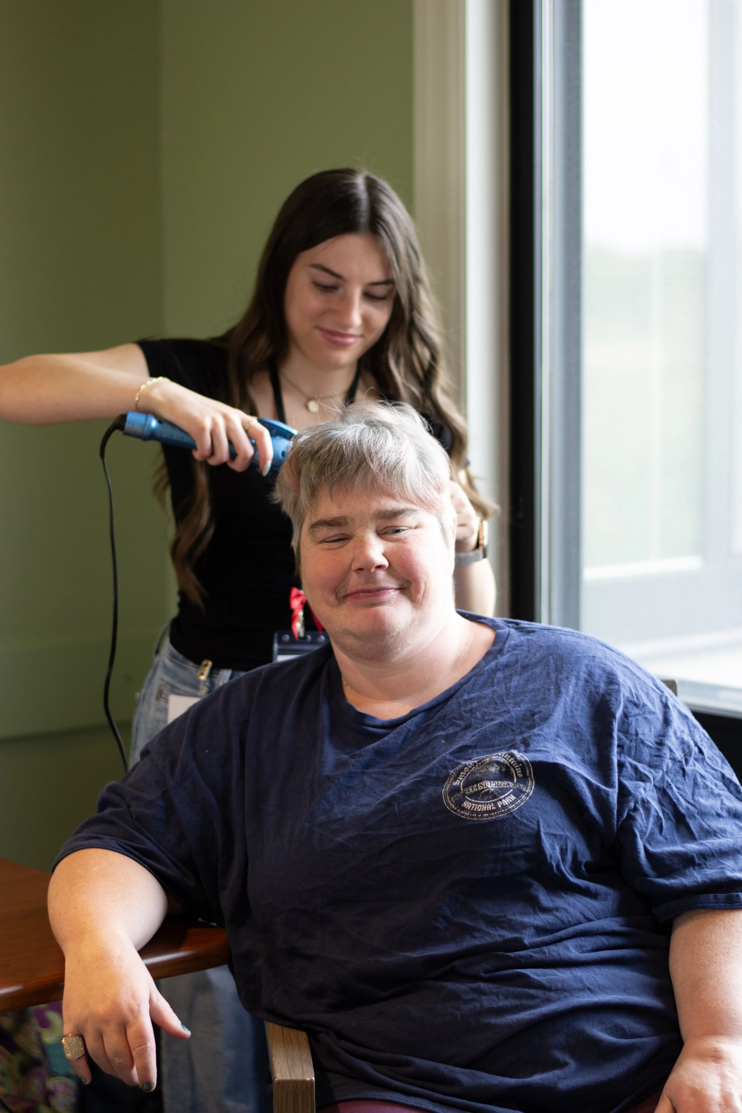 A young woman with long brown hair, wearing a black top, is trimming a man's hair or beard with electric clippers. The man is smiling, wearing a navy T-shirt, sitting on a wooden chair, near a large window with natural light.