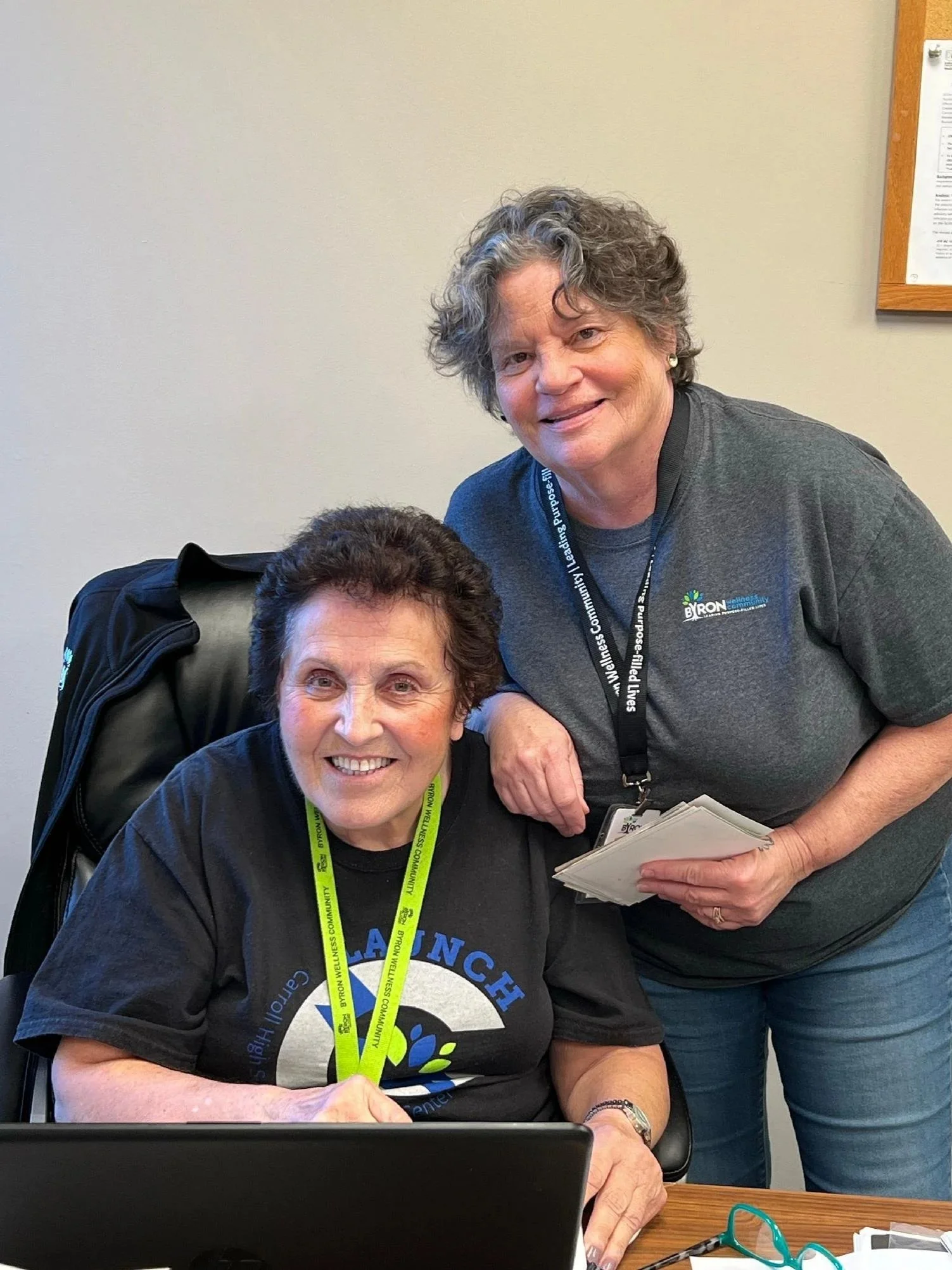 Two women in a room, one sitting at a desk with a computer, smiling, and another standing beside her, holding a notebook and smiling. Both are wearing lanyards and casual clothes.