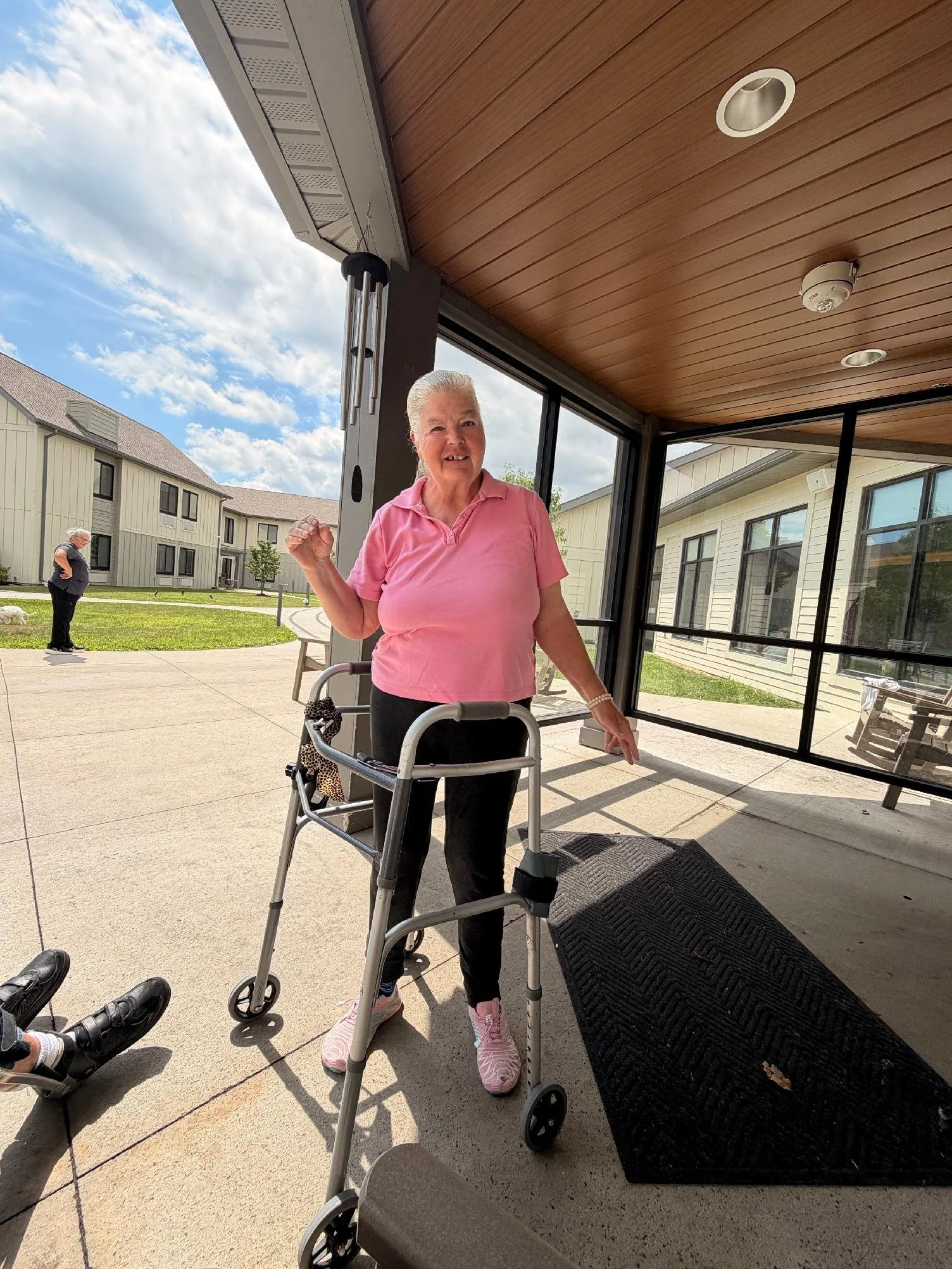 Older woman with white hair in a pink shirt standing with a walker outside of a building, smiling at the camera. In the background, another person with white hair and a dark shirt is walking on a grassy area. The setting appears to be a residential c