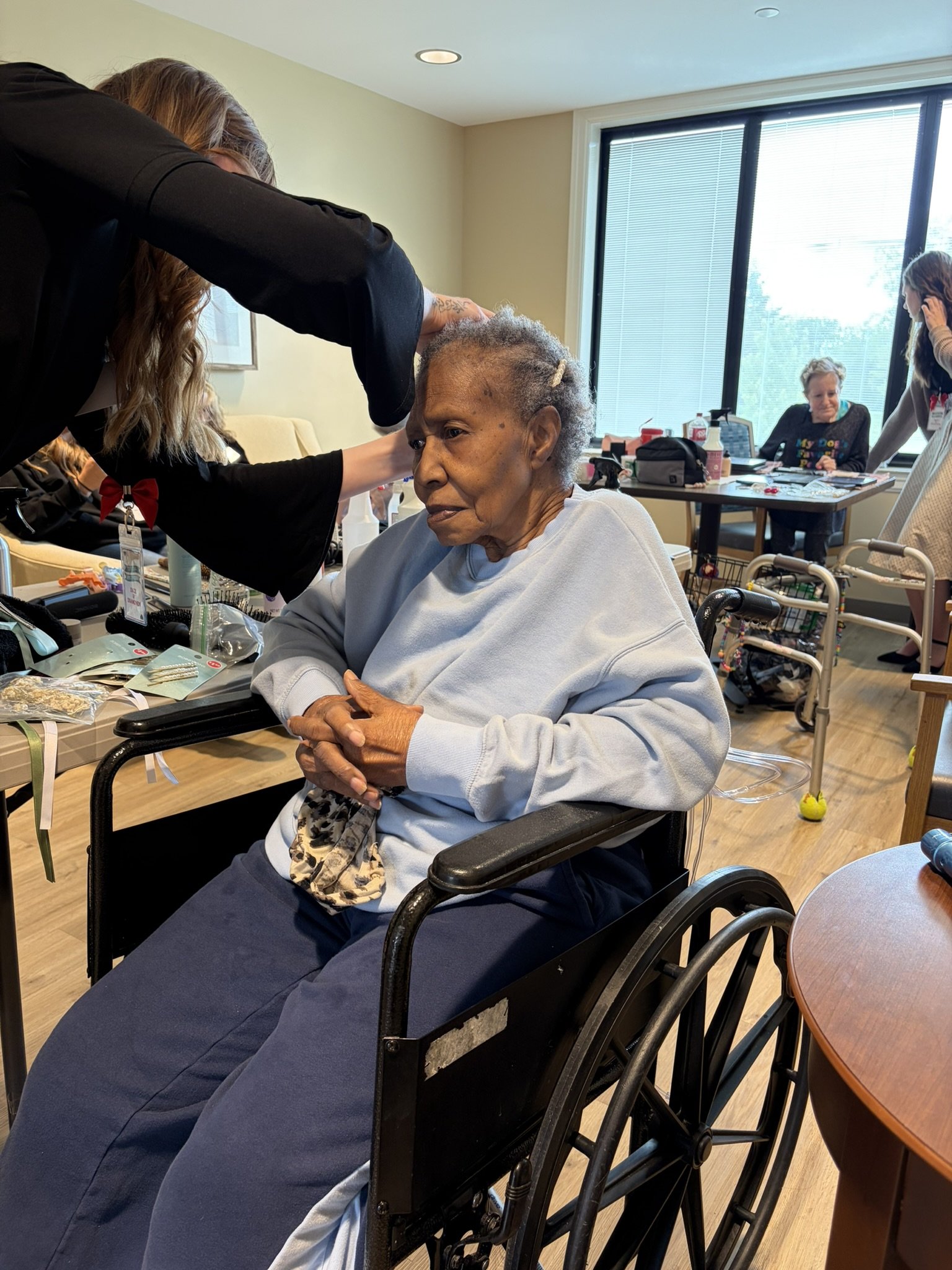 An elderly woman with gray hair in a wheelchair getting her hair styled by a woman in black attire in a room with large windows. There are people and various items on tables in the background.