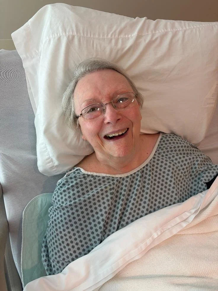 An elderly woman lying in a hospital bed, smiling and wearing glasses, dressed in a hospital gown.