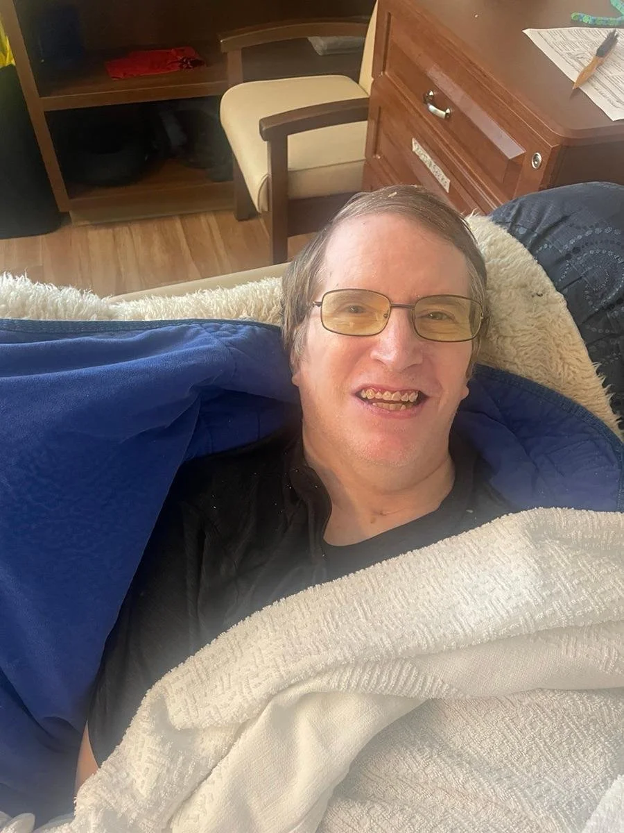 A man lying on a bed, smiling at the camera, with glasses, wearing a dark shirt and covered with a white blanket, in a room with wooden furniture.