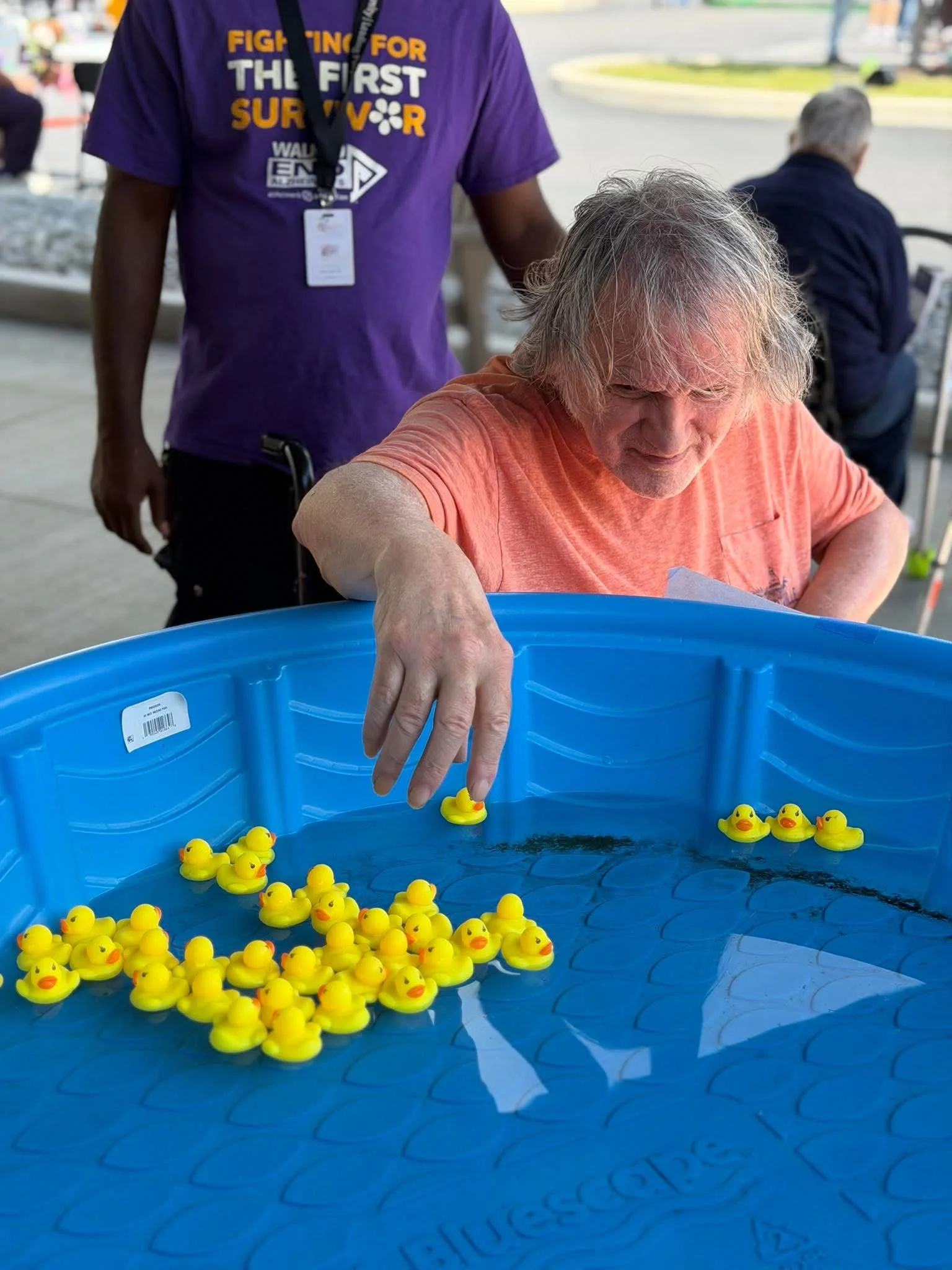 Person with gray hair in an orange shirt playing a duck fishing game at an outdoor event with plastic ducks floating in a blue water tank.