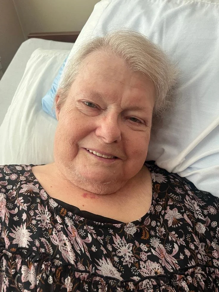A smiling elderly woman lying in a hospital bed, wearing a black floral blouse, with a pillow behind her head.