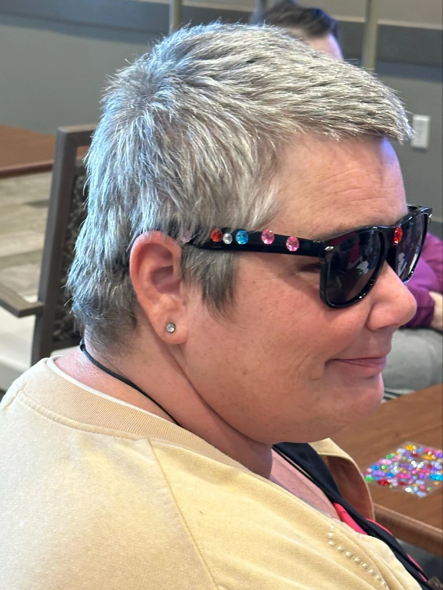 Profile of a woman with short, gray hair, wearing sunglasses with colorful rhinestones, and earrings, sitting at a table with small beads or sequins in front of her.