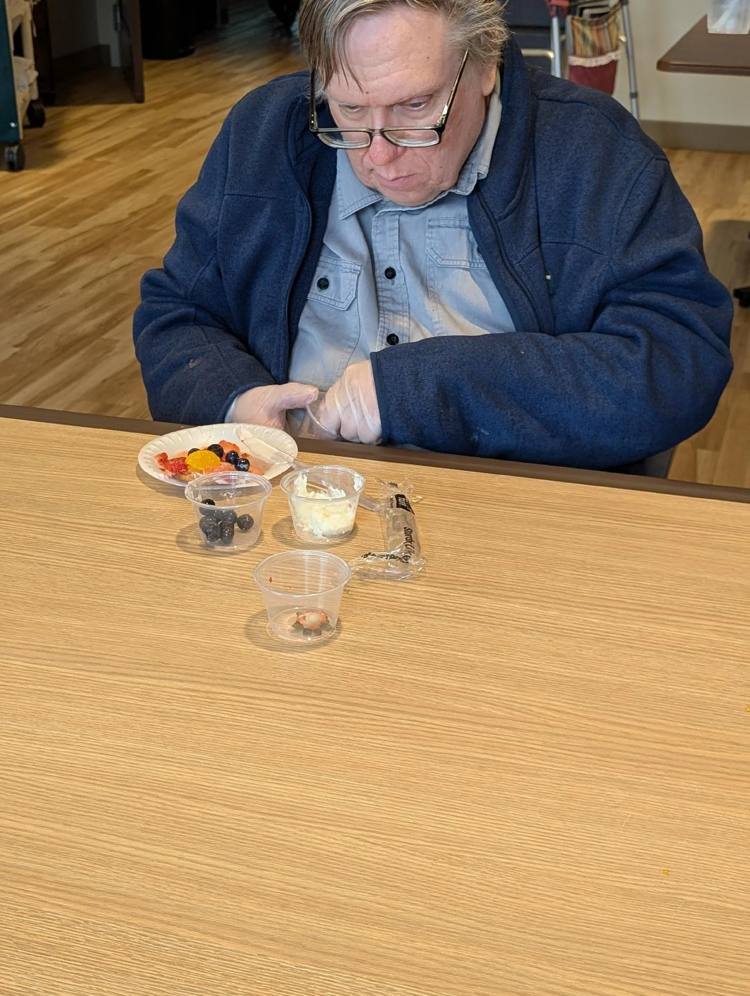 An older man with glasses wearing a blue jacket and light-colored shirt sitting at a table, examining small cups of food and fruit. The table holds small containers with blueberries, a slice of cake, and fruit.