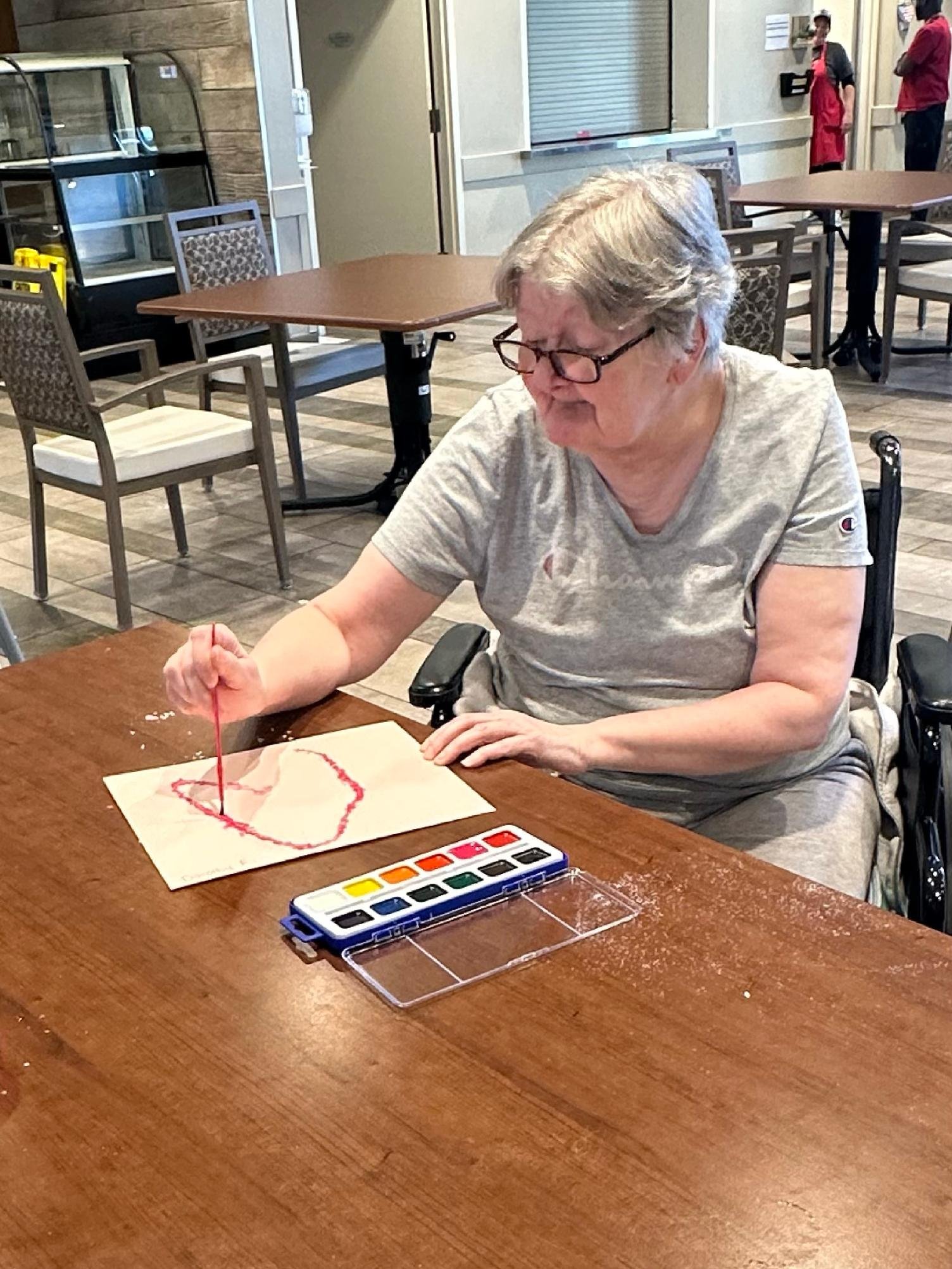 An older woman in a wheelchair making a red heart with yarn on paper at a wooden table, with watercolor paints nearby, in a casual restaurant or cafe setting.