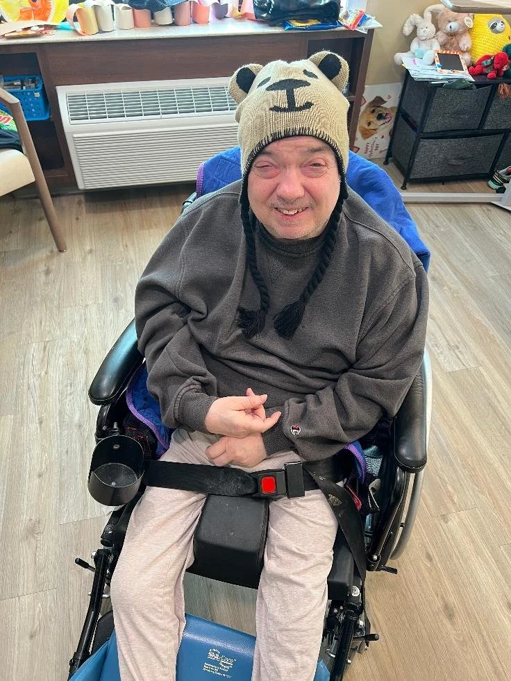 A smiling man in a wheelchair wearing a bear hat with ear details, a gray sweatshirt, and beige pants, inside a room with toys and decorations in the background.