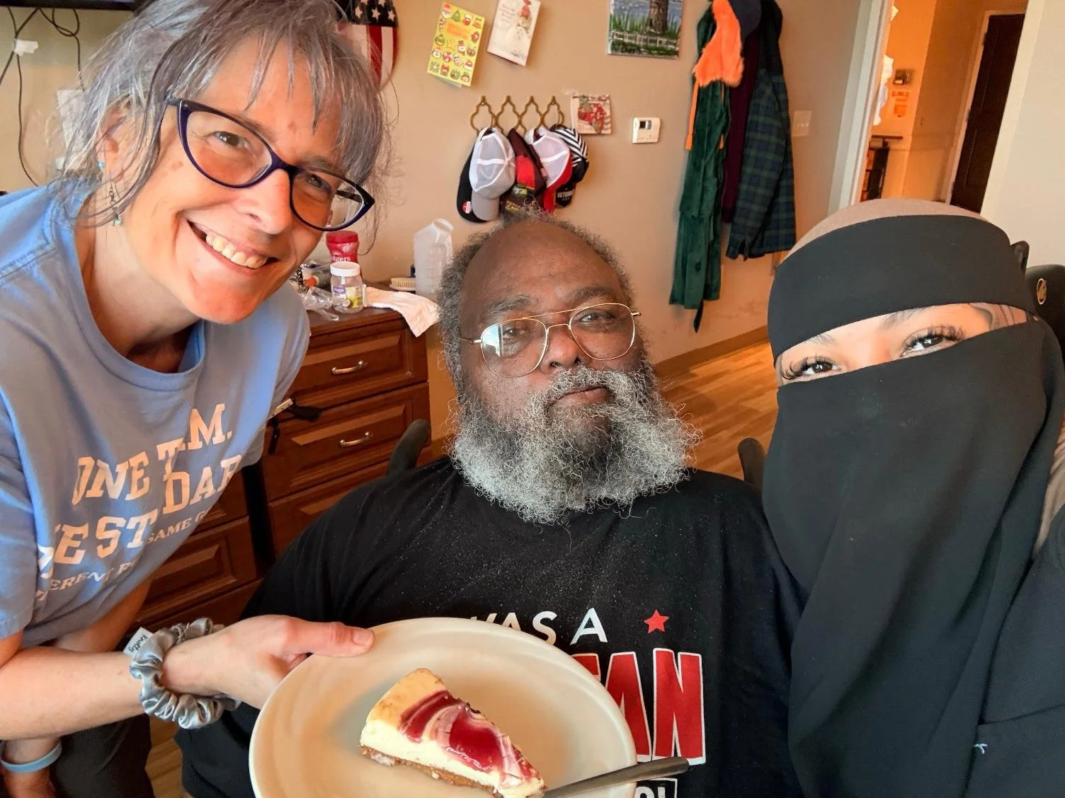 Three people posing together in a kitchen, with one holding a plate of cheesecake topped with red sauce.