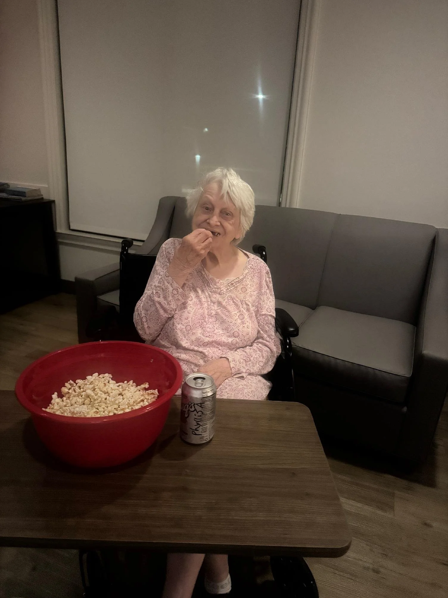 An elderly woman with white hair sitting on a black chair in a living room. She is wearing pink pajamas and eating popcorn. On the table in front of her is a large red bowl of popcorn, a can of Sprite soda, and a soda can labeled PLYO. Behind her is 