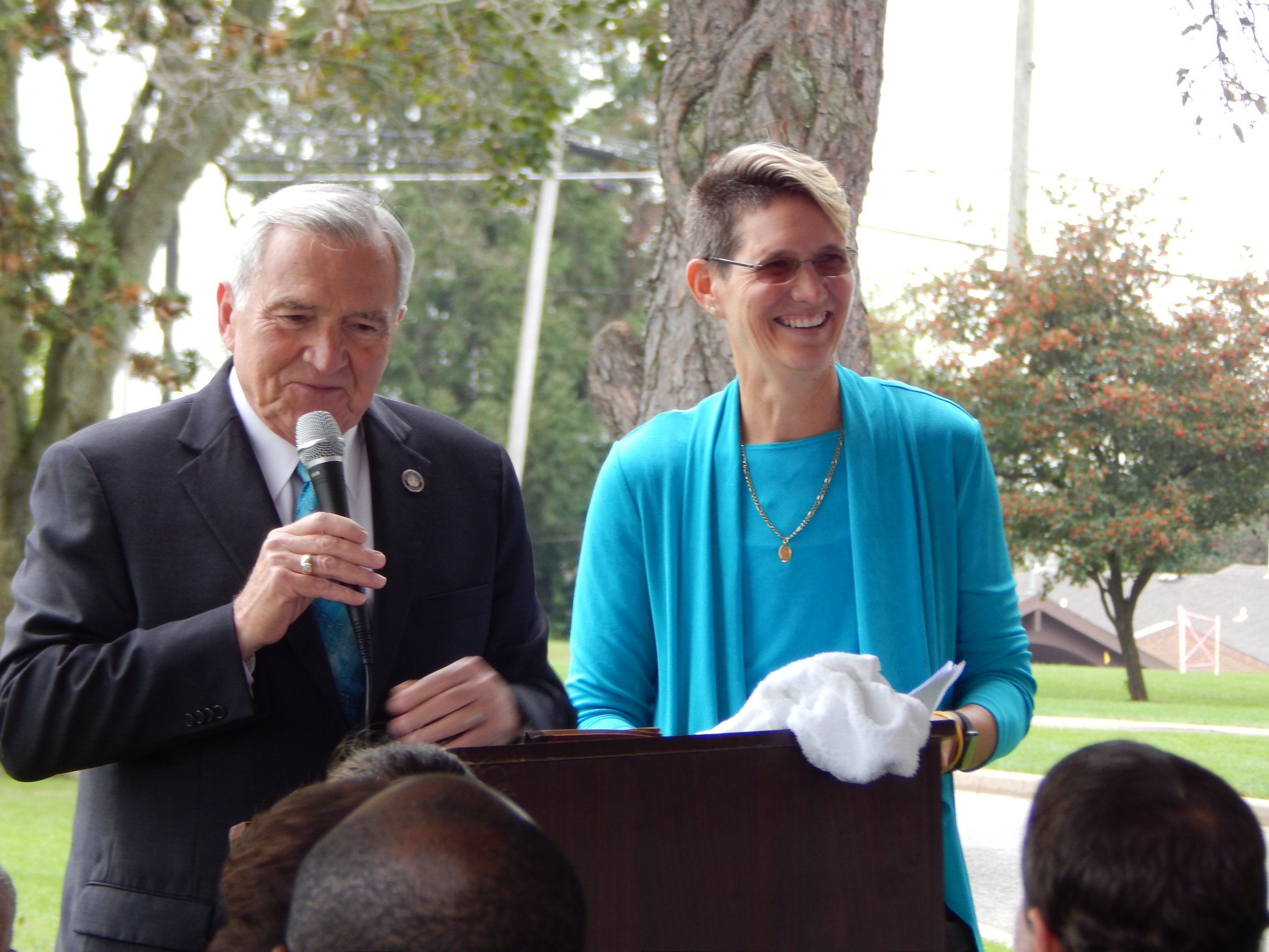 Two adults speaking at an outdoor event. The man on the left is holding a microphone and appears to be reading or speaking. The woman on the right is smiling and looking towards the audience. They are standing beneath trees in a park or garden setting.