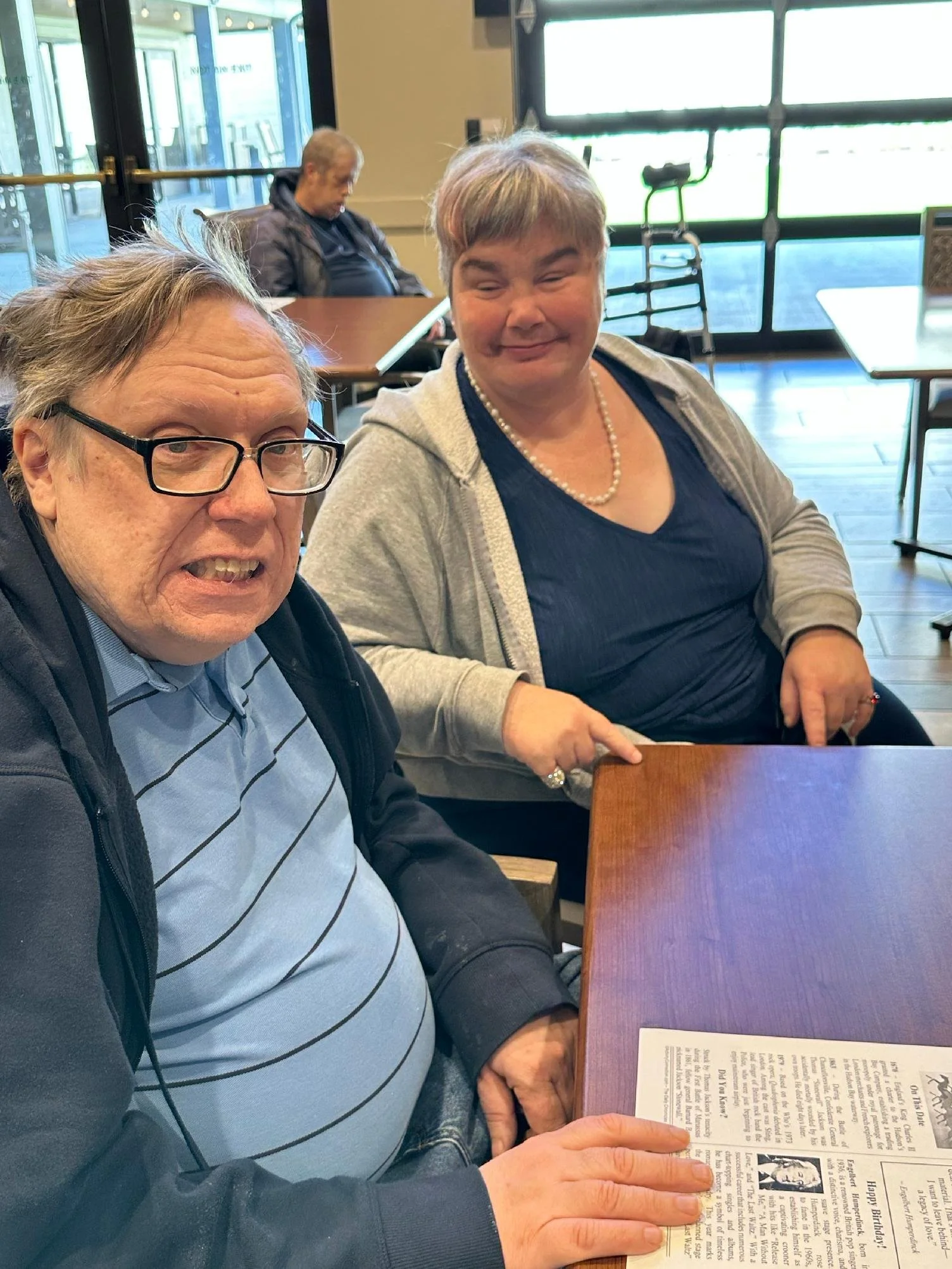 Two people sitting at a wooden table in a restaurant, looking at a newspaper, with a third person asleep in the background. The man on the left is wearing glasses and a striped polo shirt, and the woman on the right has short hair, a pearl necklace, 