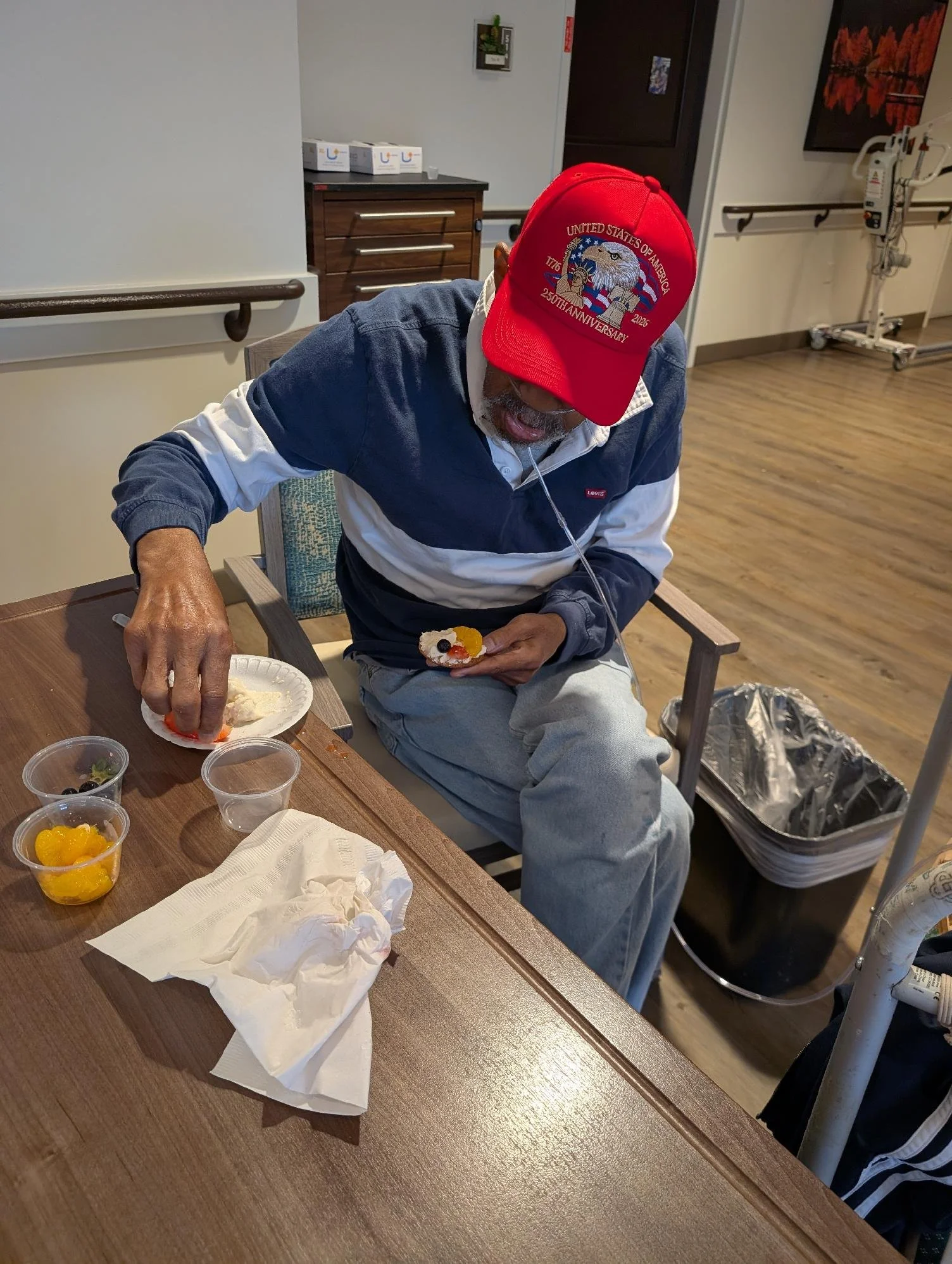 An elderly man with a beard wearing a red baseball cap and a blue and white jacket sitting at a table, eating a dessert with fruit and whipped cream, in what appears to be a hospital or care facility dining area.