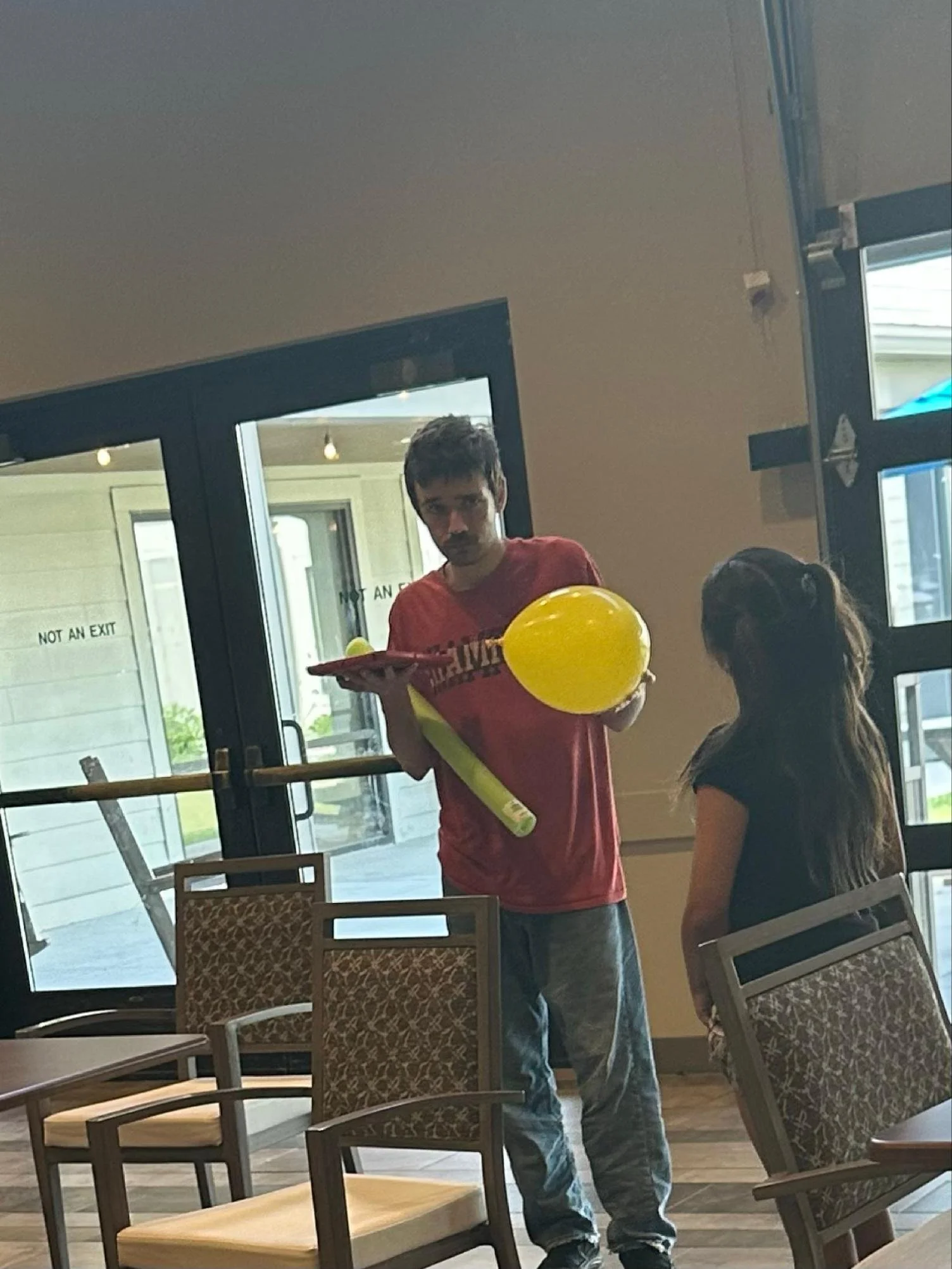 A man with dark hair and a mustache wearing a red shirt and jeans holding yellow balloons and a tray in a restaurant or cafe, with a woman with long hair standing near him, behind a glass door with signs that read ‘Not An Exit’.