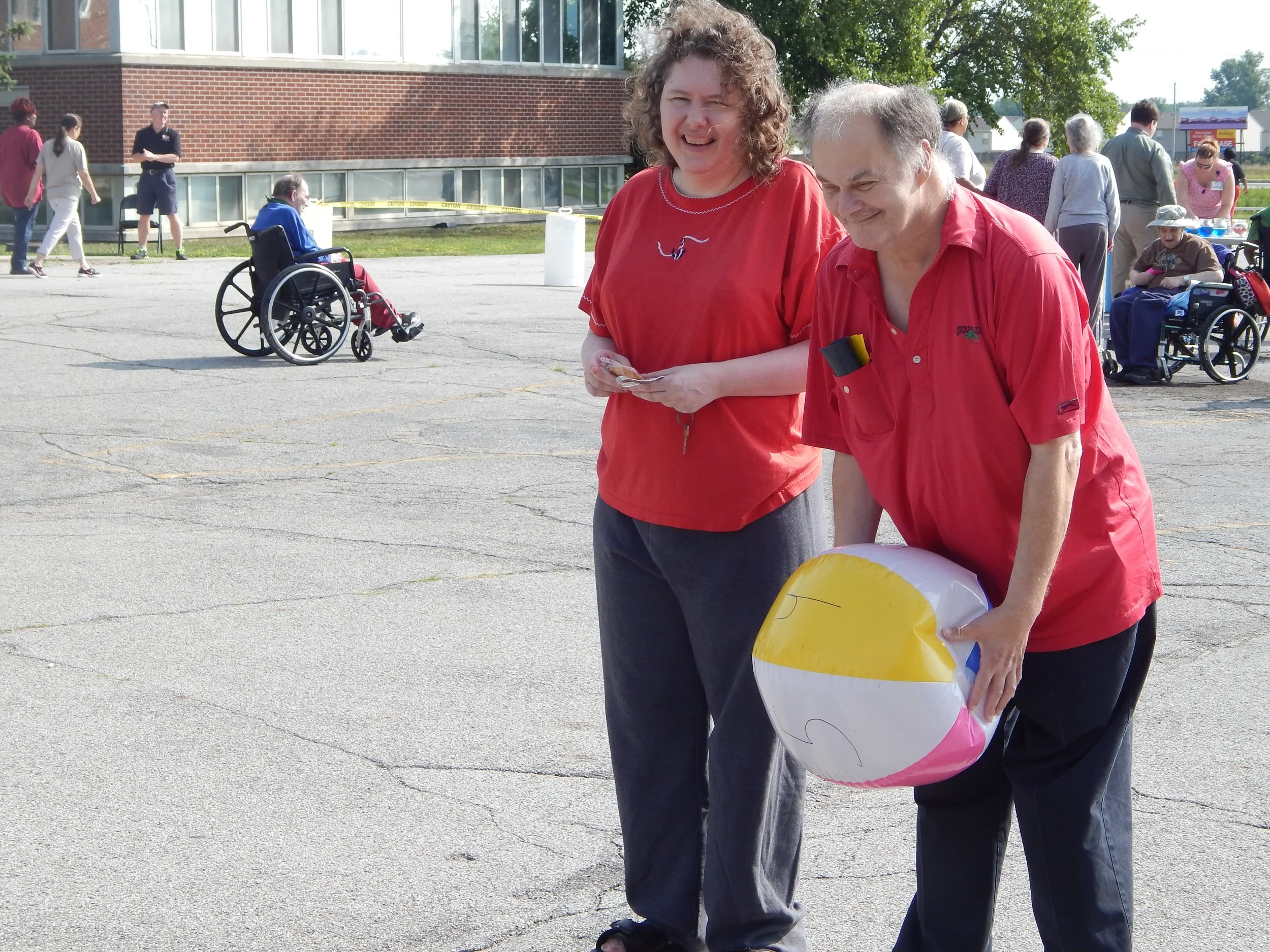 Two women stand close together outdoors during the daytime, smiling and holding balloons, with other people and a building in the background.