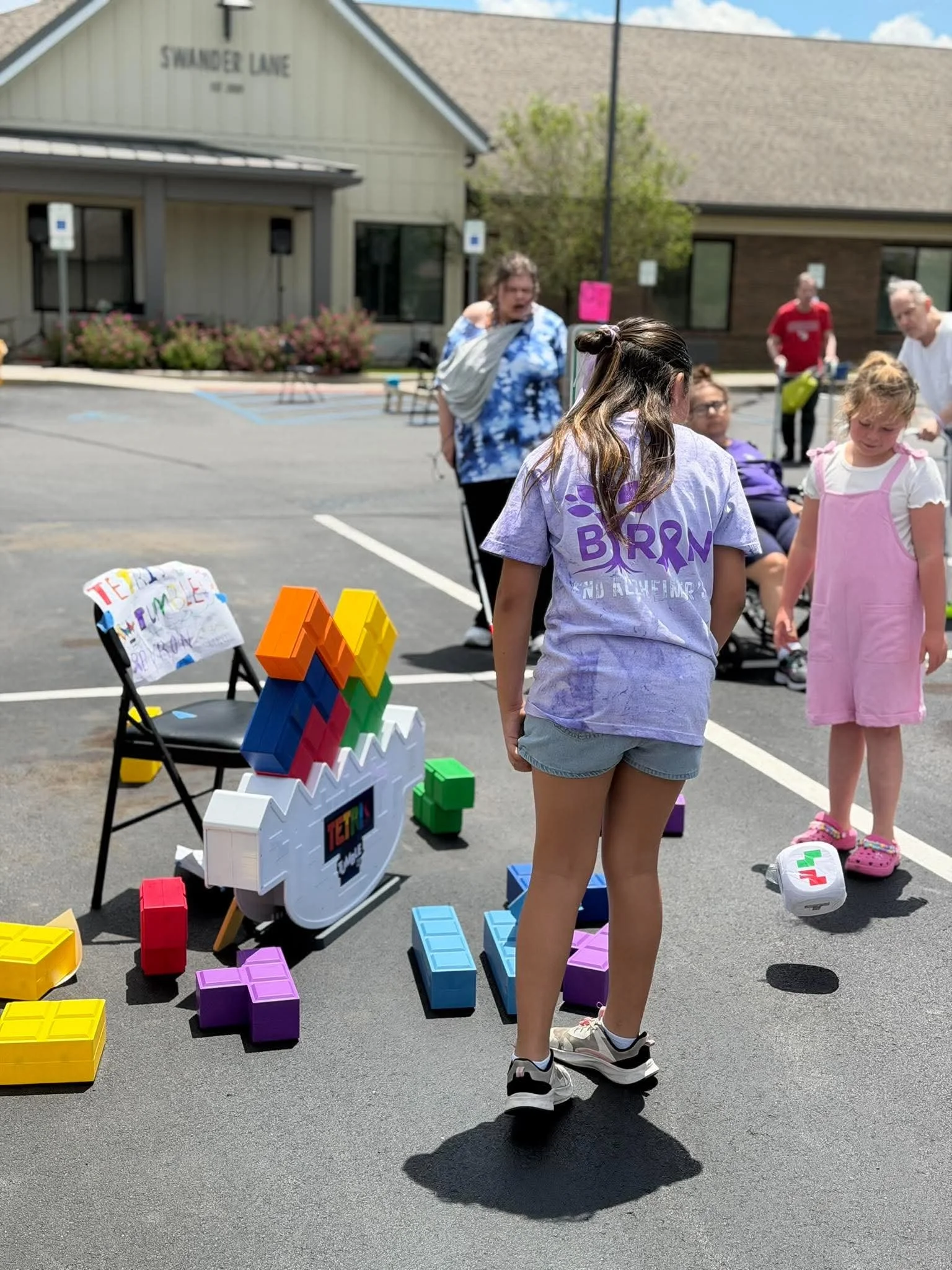 People gathered outside in a parking lot with large building in background. Various colorful building blocks and a small structure made of blocks are on the ground.