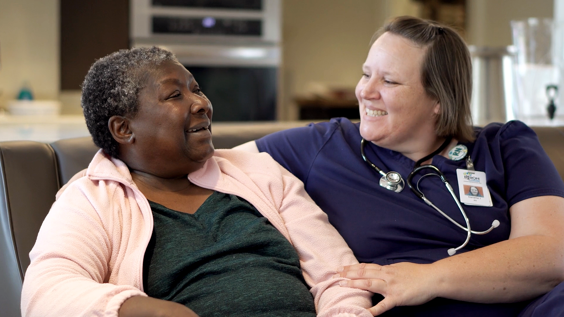 A healthcare professional, wearing a navy blue uniform with a stethoscope and a name tag, smiling and sitting beside an older woman with short, curly gray hair, wearing a pink jacket and dark shirt, in a healthcare or hospital setting.