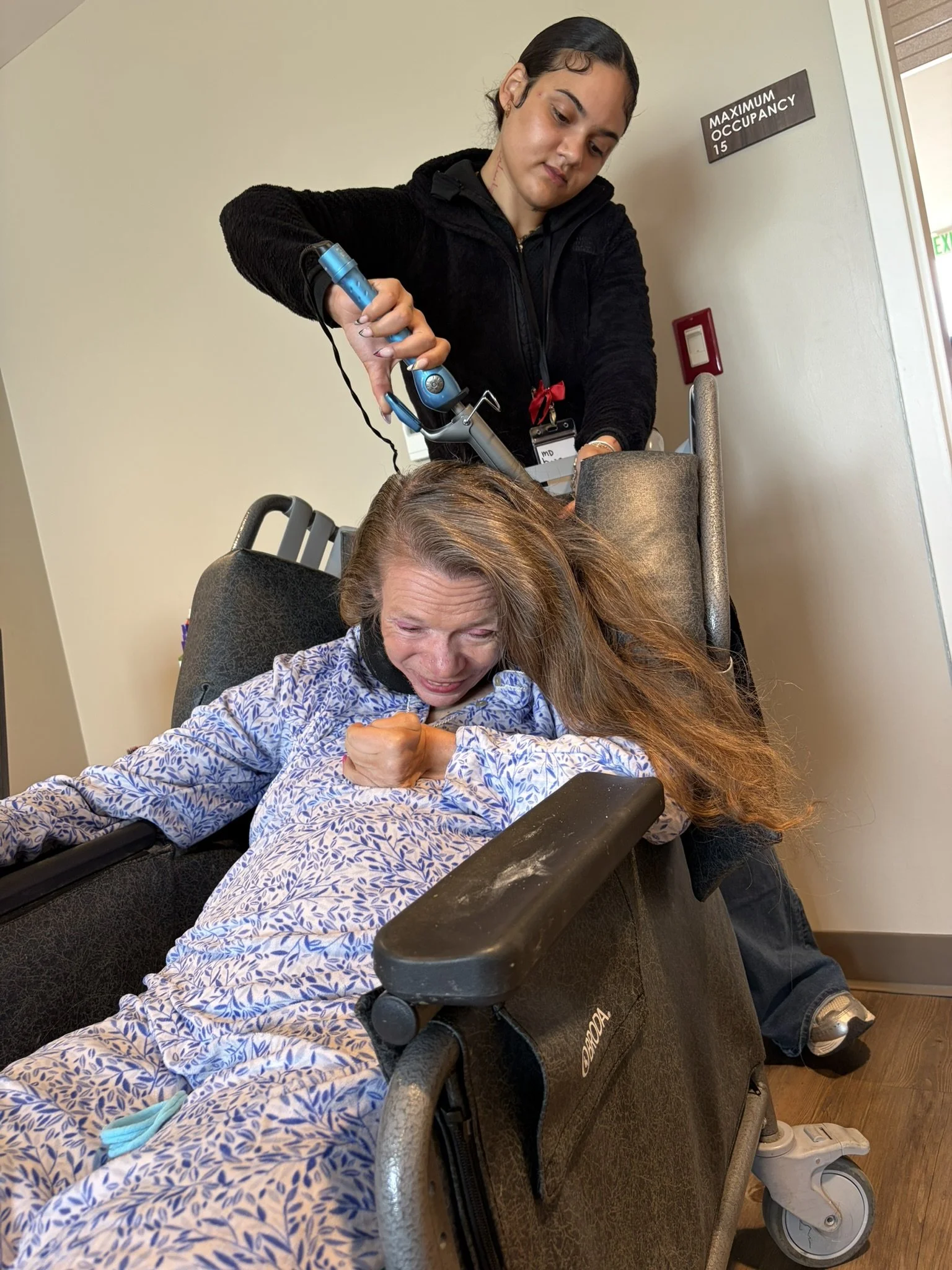 A young woman with long hair is sitting in a wheelchair, laughing and clenching her fist. A nurse is standing behind her, placing a portable oxygen concentrator's tube into her nose. The woman is wearing a patterned gown. The setting appears to be a 