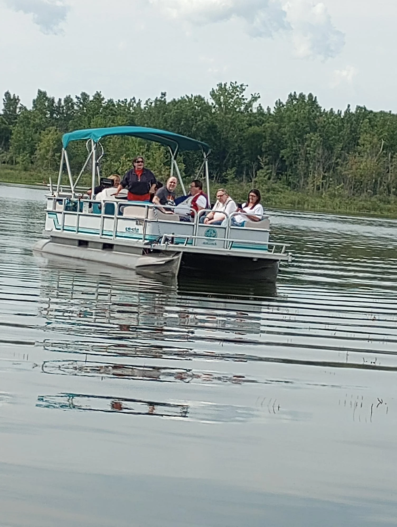 A group of people riding on a pontoon boat on a lake, surrounded by greenery and trees in the background.