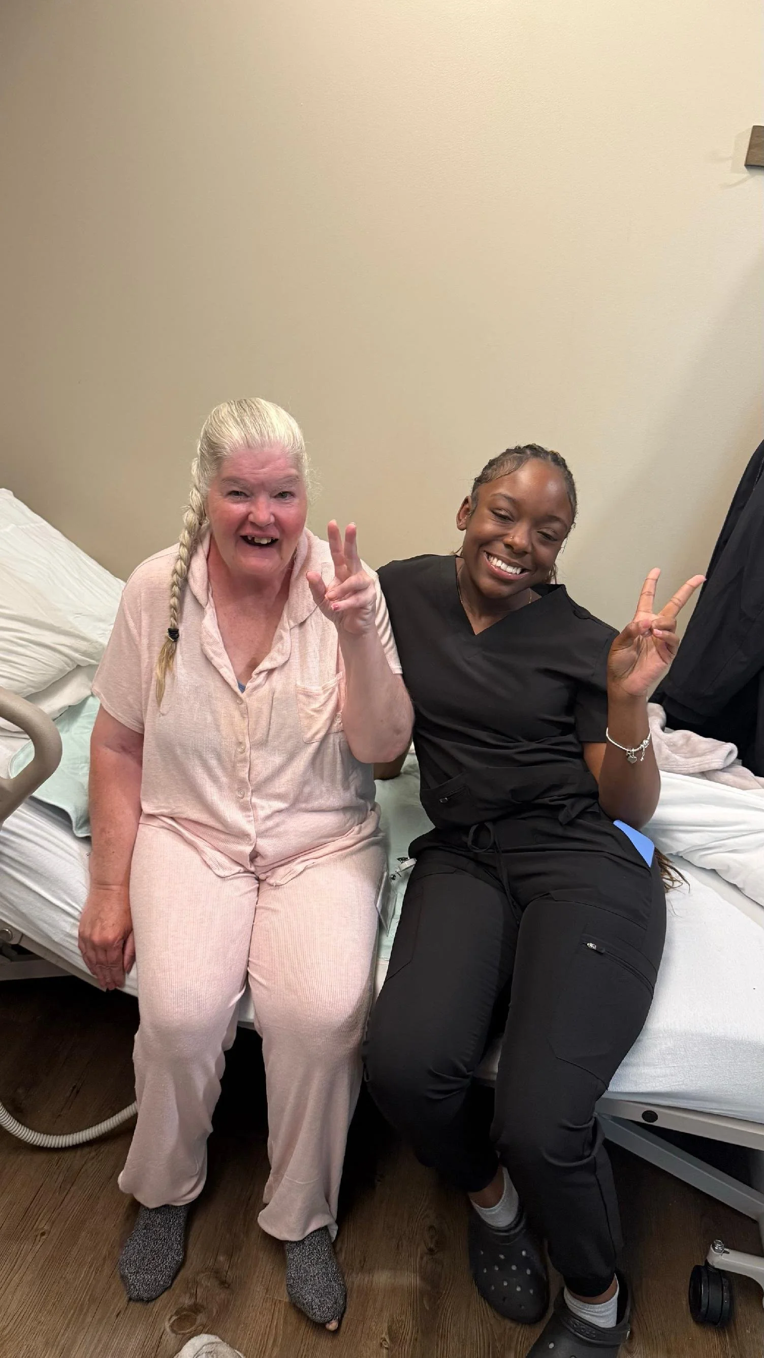 Two women sitting on a hospital bed, smiling and making peace signs. The older woman on the left has light skin, white hair in a braid, and is wearing light pink pajamas. The younger woman on the right has dark skin, dark braided hair, and is dressed