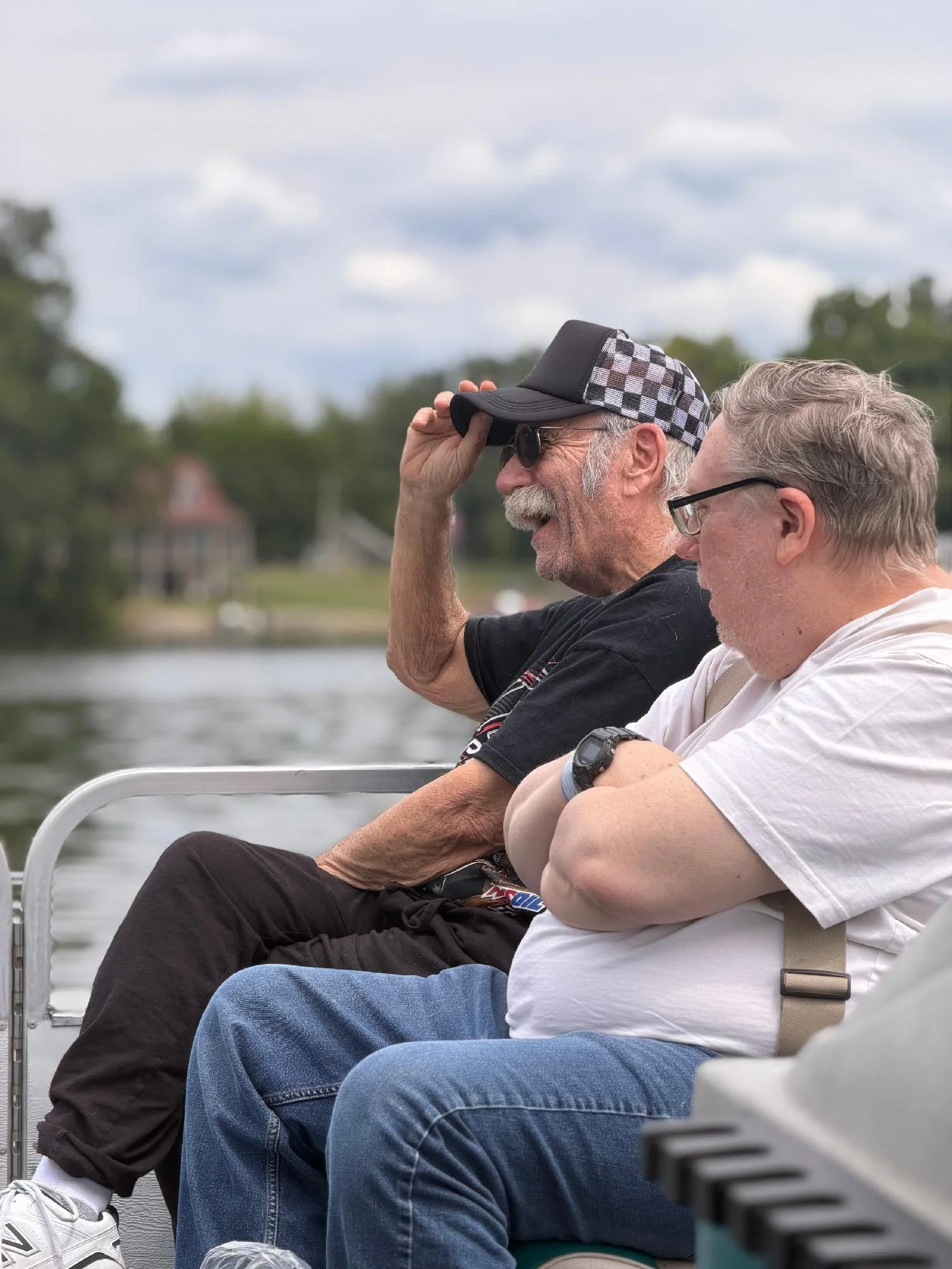 Two older men sitting outdoors on a boat or dock near a body of water, one with a checkered cap and sunglasses, the other with glasses, both looking out at the water under a cloudy sky.