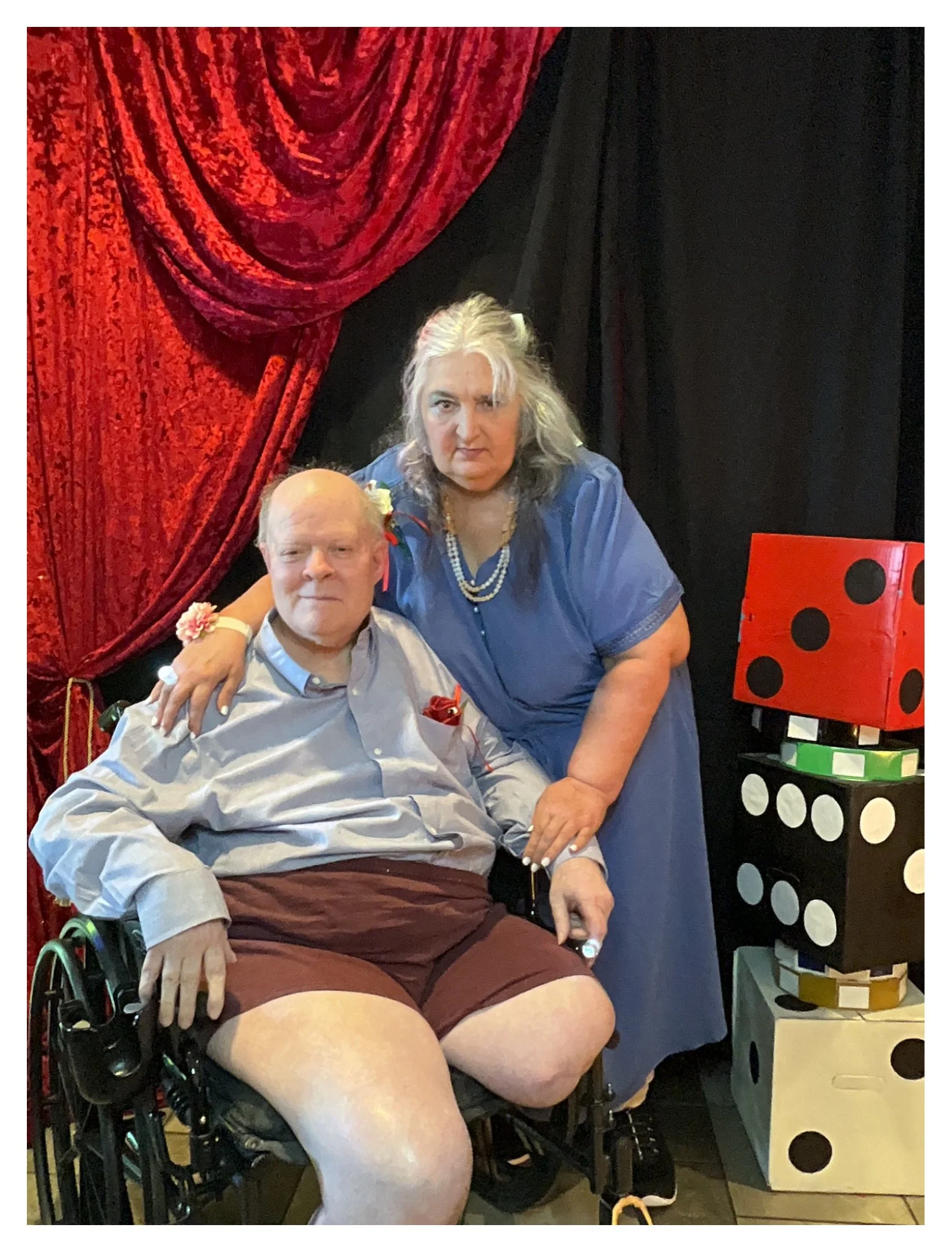 A man in a wheelchair with a blonde woman standing next to him, both with serious expressions, surrounded by red and black curtains and a large set of dice stacked on a table.