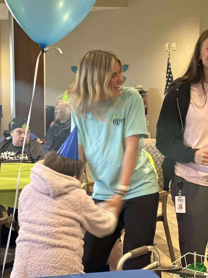A woman in a light blue shirt is smiling and holding hands with a young girl in a pink hoodie and blue party hat. The girl is reaching up and holding the woman's hand. There are birthday balloons and party hats, indicating a celebration.