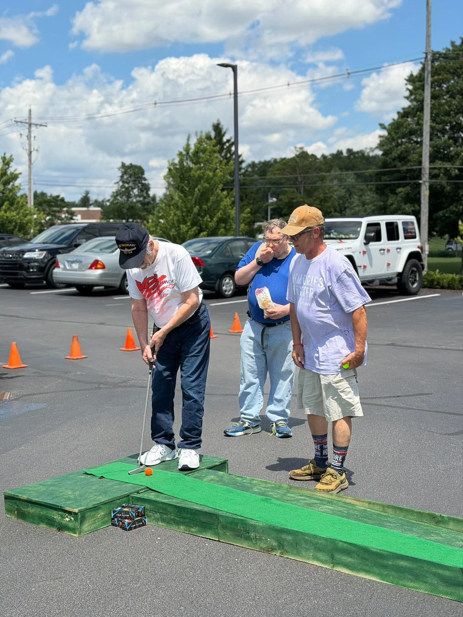 Three men play mini golf outdoors on a sunny day with a parking lot, trees, and a blue sky in the background. One man is lining up a shot with a putter while the others watch.