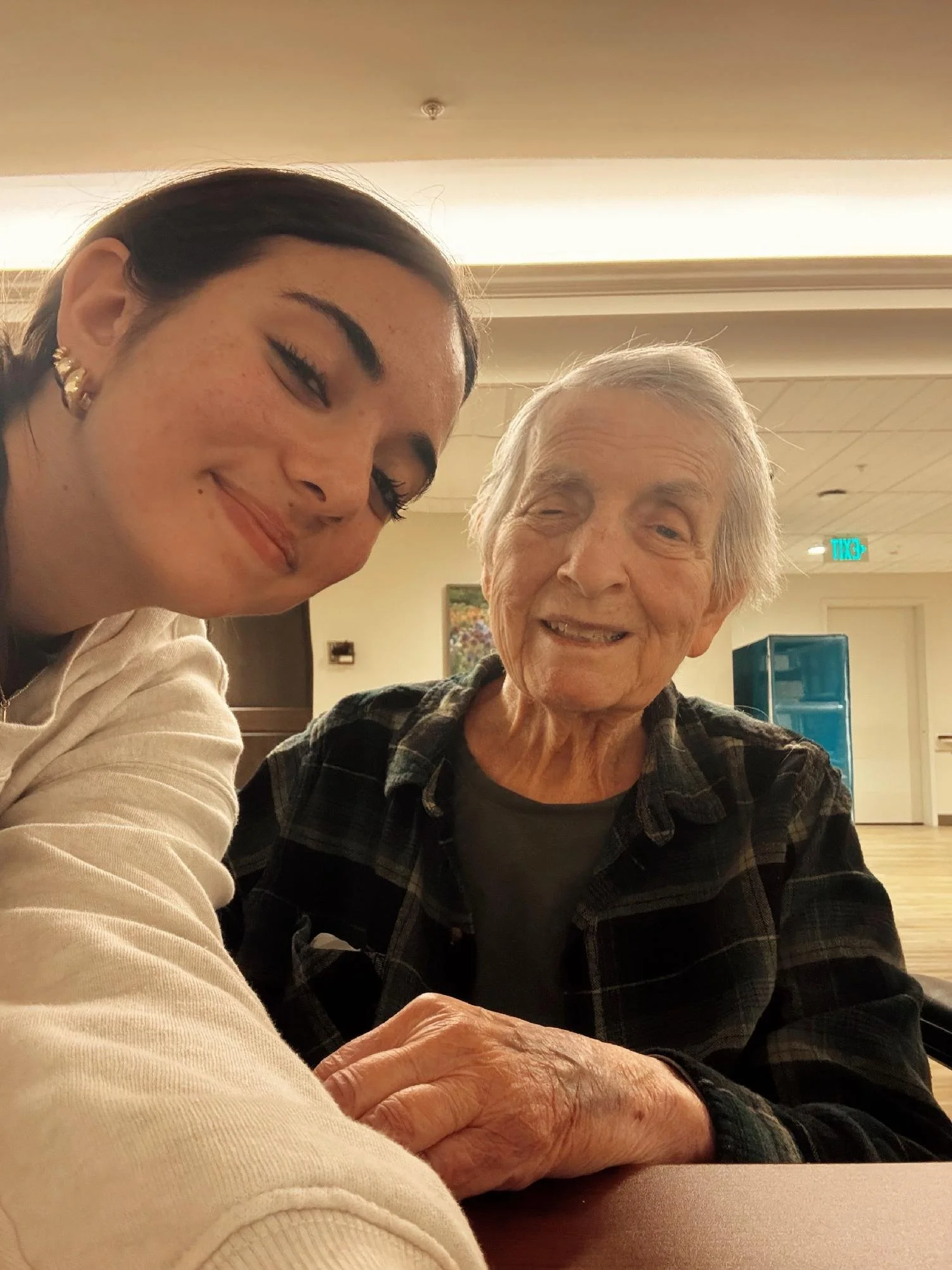 A young woman and an elderly woman smiling for a selfie, with the elderly woman wearing a plaid shirt and the young woman wearing earrings and a light-colored top.