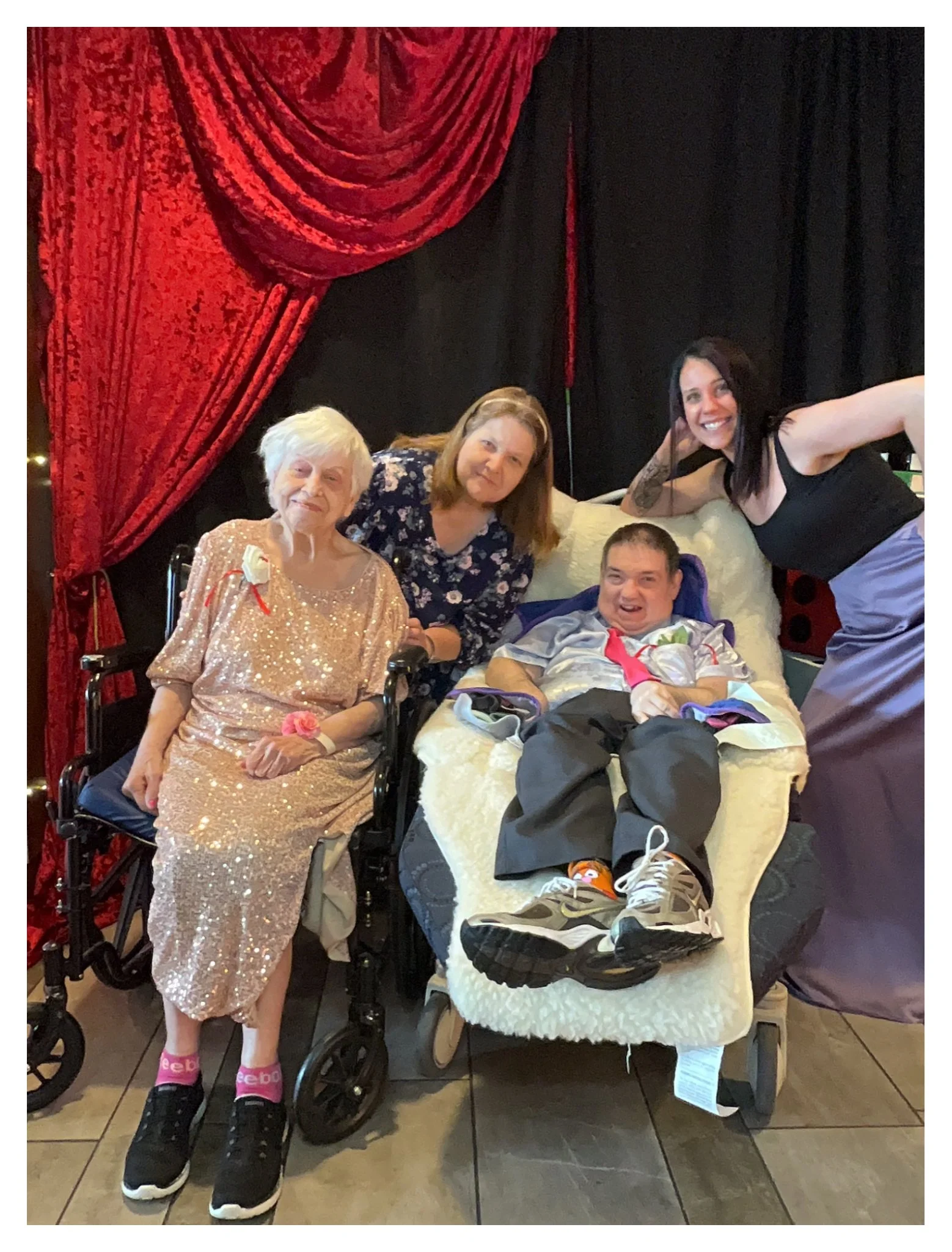 Four people, including a young man in a wheelchair with a woman beside him, a nurse or caregiver and an elderly woman, gather together in a room with curtains and black drapes in the background. They are smiling and appear to be celebrating a special