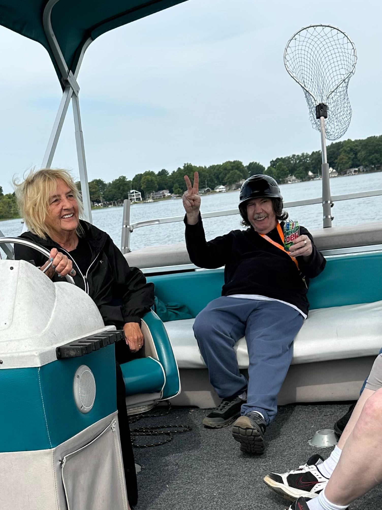 Two smiling people on a boat with a lake and trees in the background. One person is making a peace sign and holding a drink.