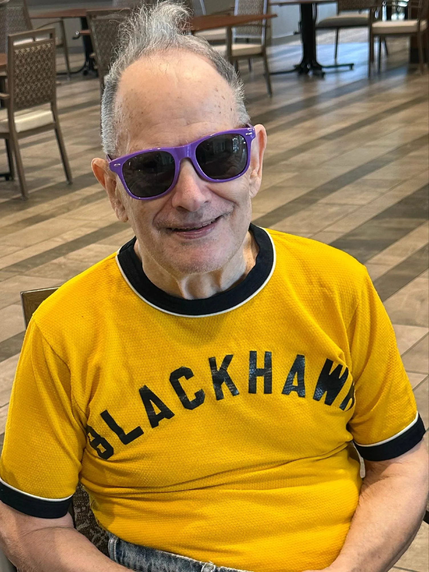 A smiling man wearing purple sunglasses and a bright yellow T-shirt with black text, sitting in a restaurant with wooden floors and chairs in the background.