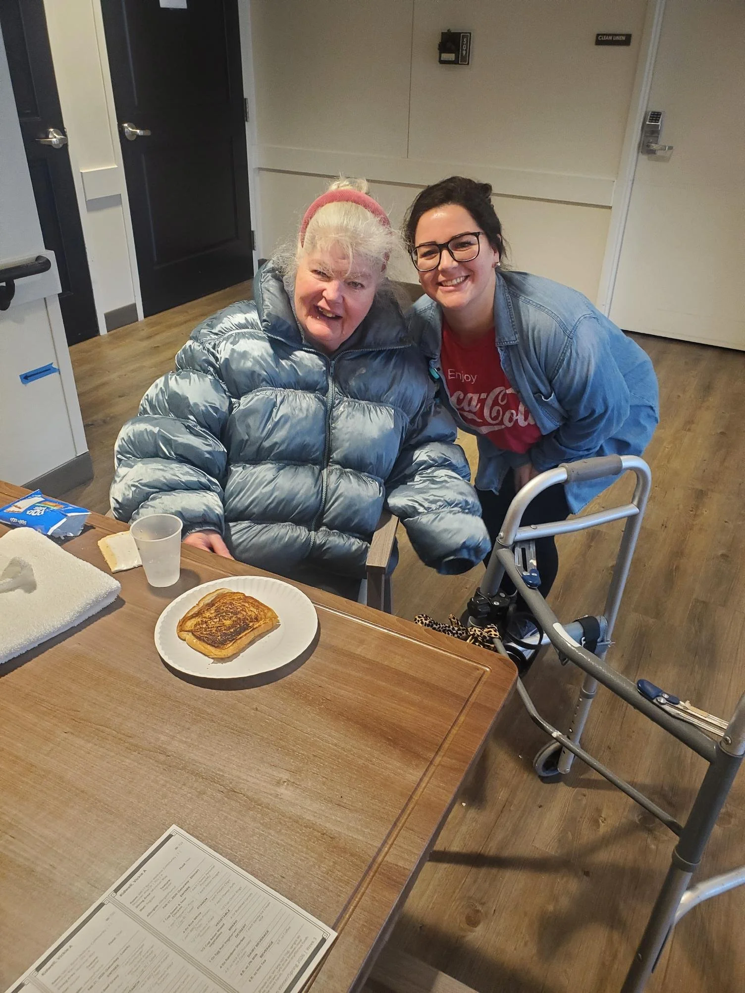 A woman with glasses and a younger woman are sitting and kneeling together at a table, smiling for the camera. The older woman is seated in a wheelchair, wearing a shiny puffer jacket and a pink headband. On the table in front of them, there is a plate with toast and a small cup.