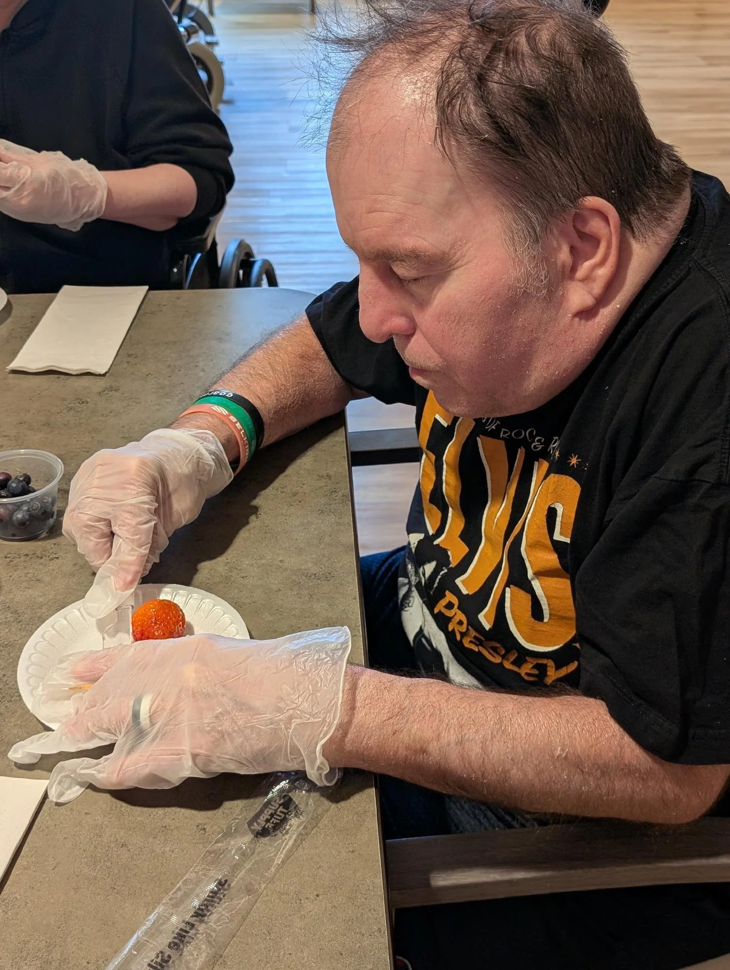 A man in a black and orange T-shirt is sitting at a table, wearing gloves, and placing a strawberry onto a paper plate that already has a strawberry and some orange slices. Another person is in the background wearing gloves, with a container of blueb