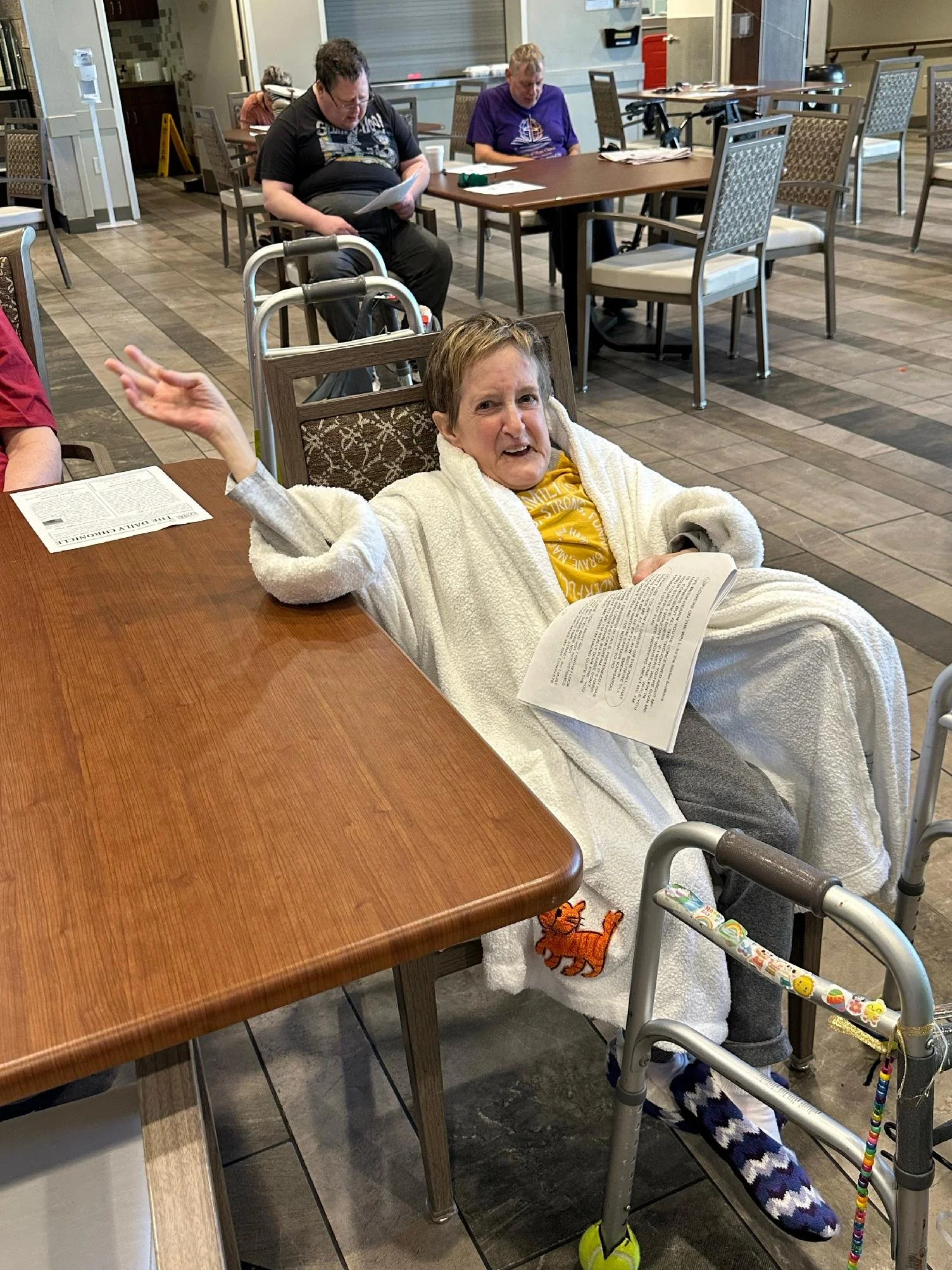 A smiling woman in a robe sitting in a wheelchair at a dining table, holding a magazine, with her feet resting on a footrest. An elderly man in a black T-shirt and dark pants sits behind her, looking at papers, while another elderly woman in a purple