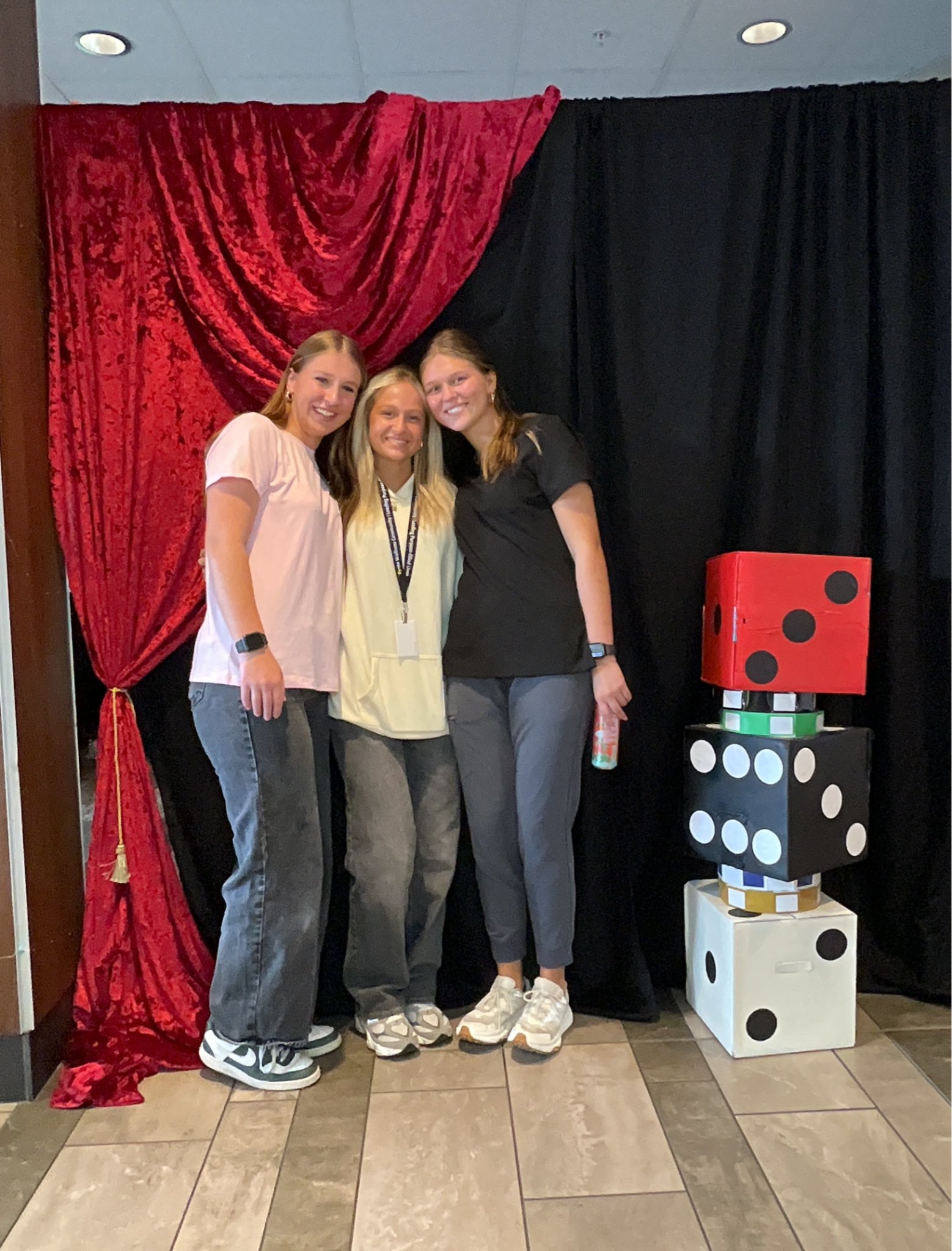 Three women standing in front of a red and black curtain with large dice decorations on the right side of the image. The women are smiling, dressed casually, and wearing sneakers.