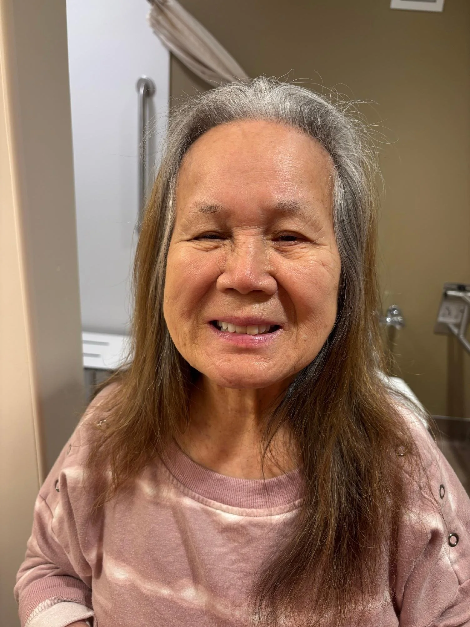 A smiling elderly woman with long gray hair and light skin, wearing a pink top, indoors with a beige wall and a sink in the background.