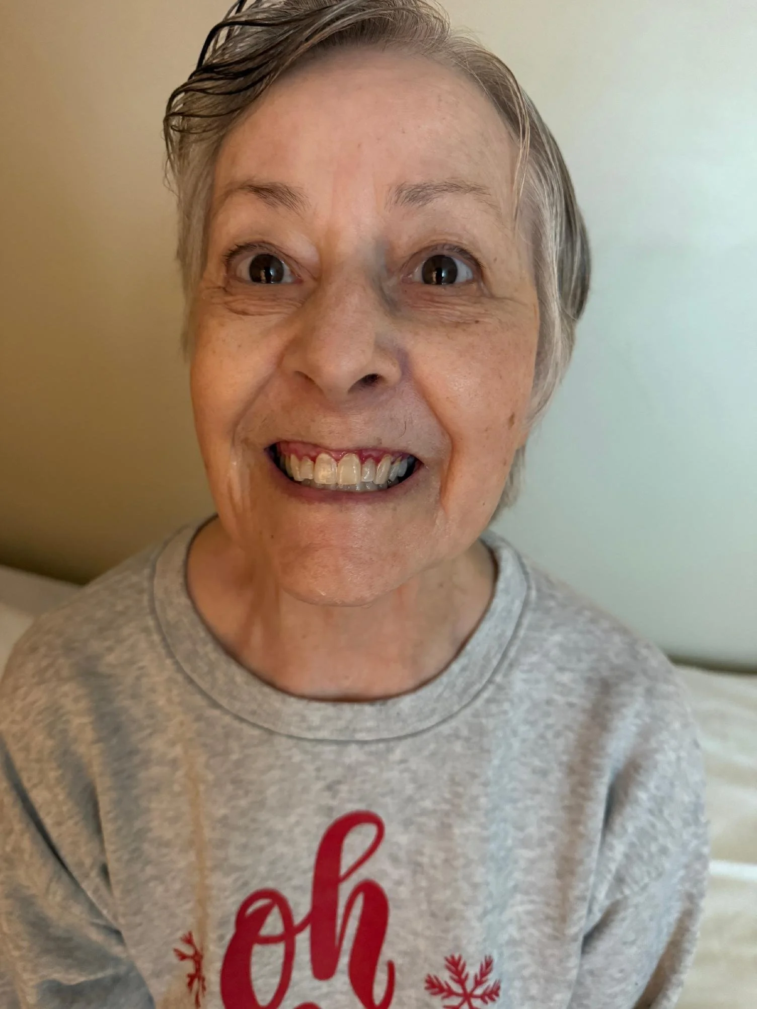 A smiling woman with short gray hair, wearing a gray sweatshirt with red lettering and snowflake designs, standing against a neutral background.