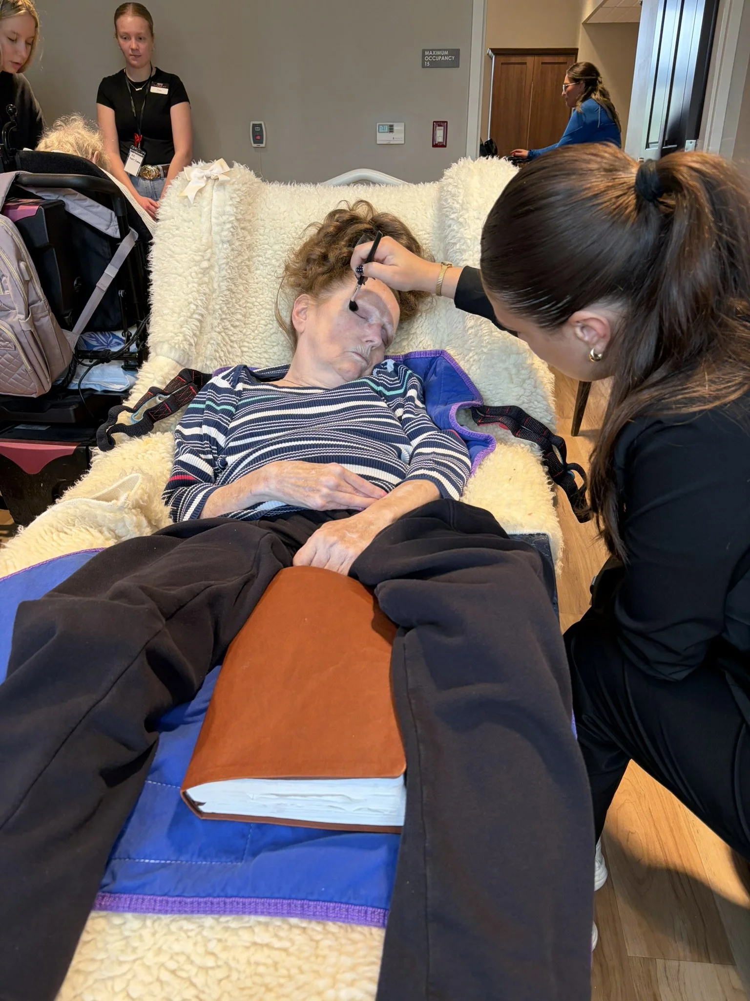 A woman with curly hair lying on a hospital bed with a fluffy cream-colored blanket, receiving makeup application from a woman with braided hair. Several people, including a young girl in a wheelchair, are present in the background.