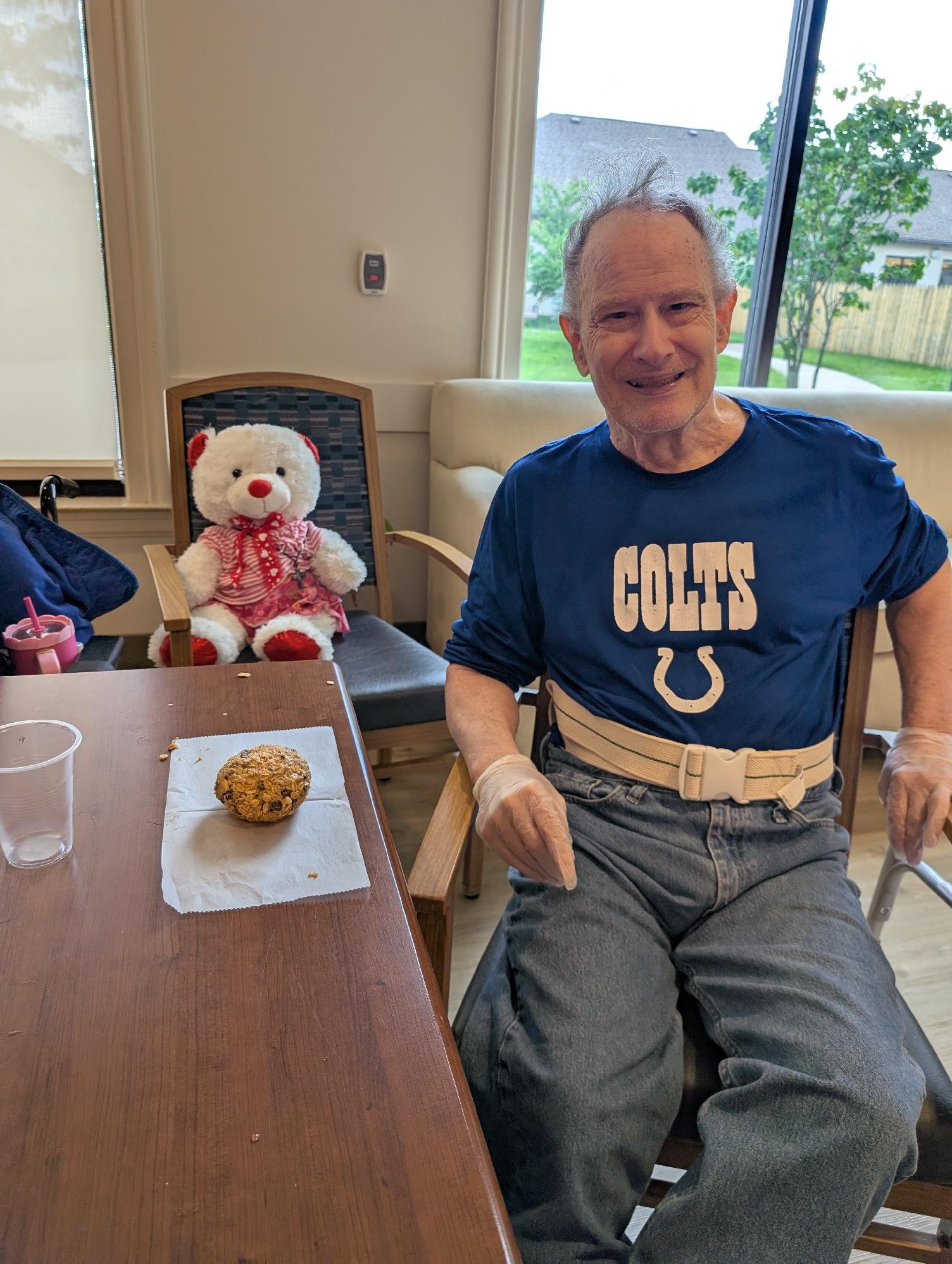 An elderly man with gray hair sitting at a table, smiling, wearing a blue T-shirt with a Colts logo, and gray pants. There is a teddy bear with a red nose and a red and white dress sitting on a chair behind him. On the table, there is a muffin, a cle